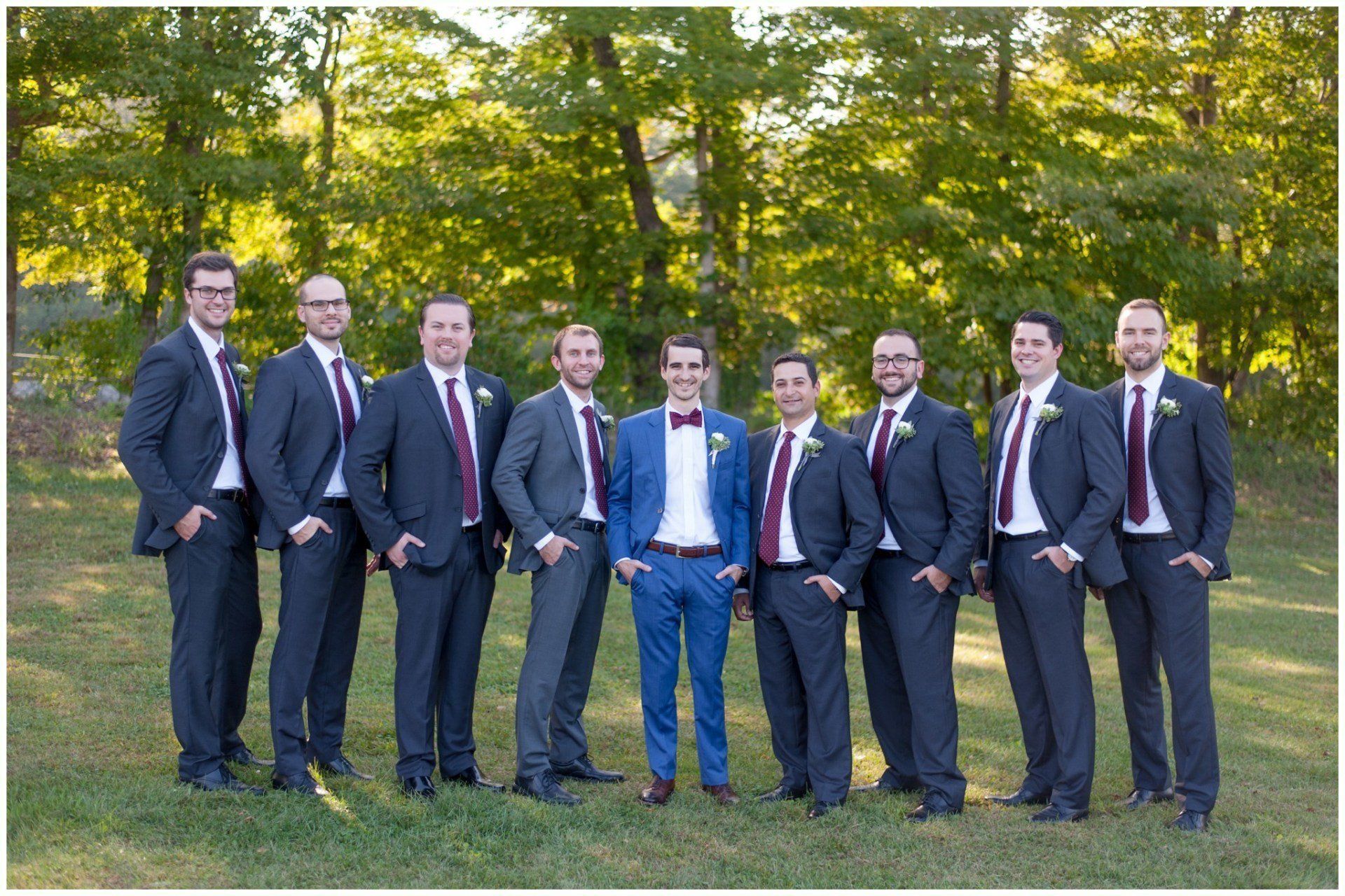 Nine men in suits stand together outdoors on grass in front of trees.