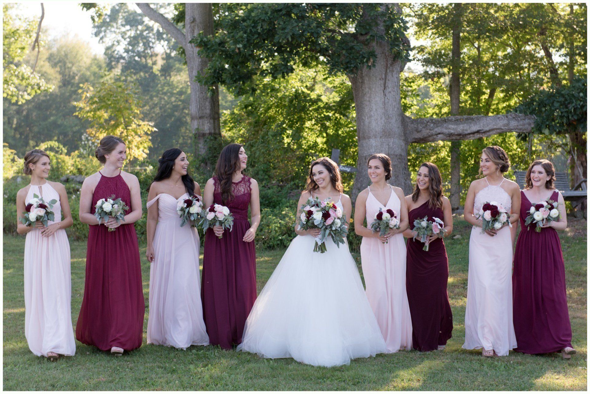 Bride and bridesmaids in white and burgundy dresses holding bouquets outdoors under trees