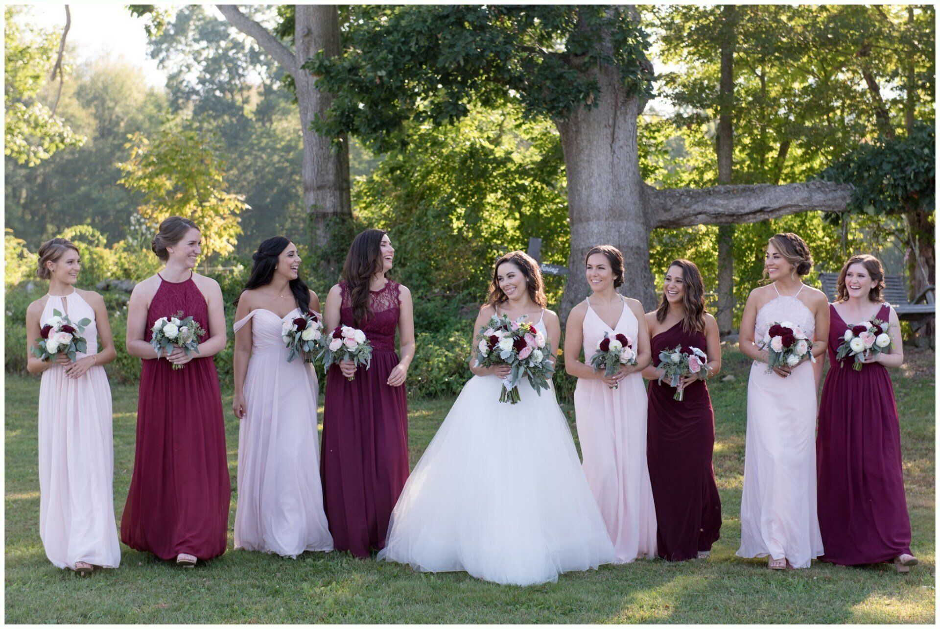 Wedding party posing outdoors in white and burgundy dresses, holding bouquets on a sunny lawn.