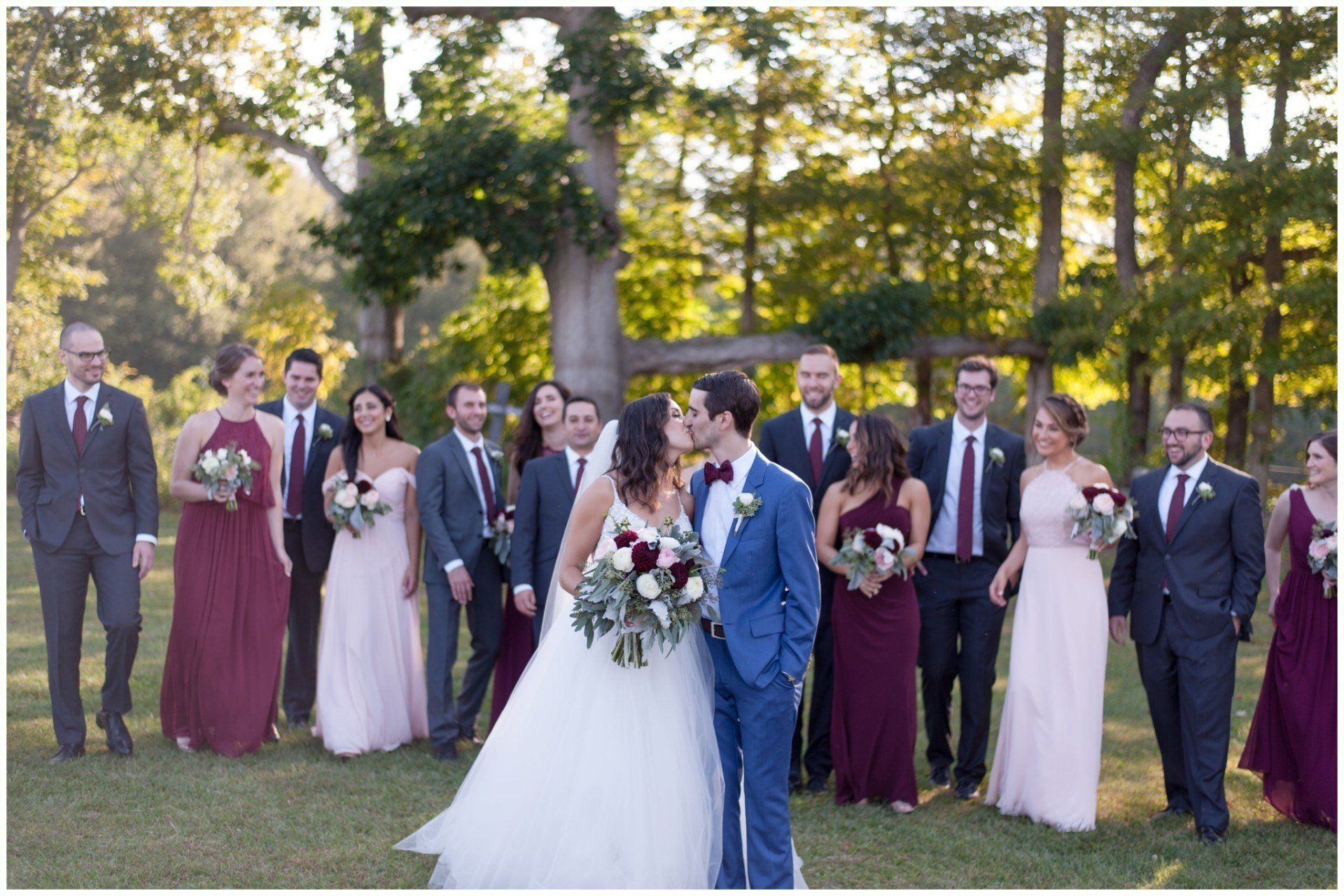 Wedding party posed outdoors in formal attire, with bride and groom centered in front of trees.