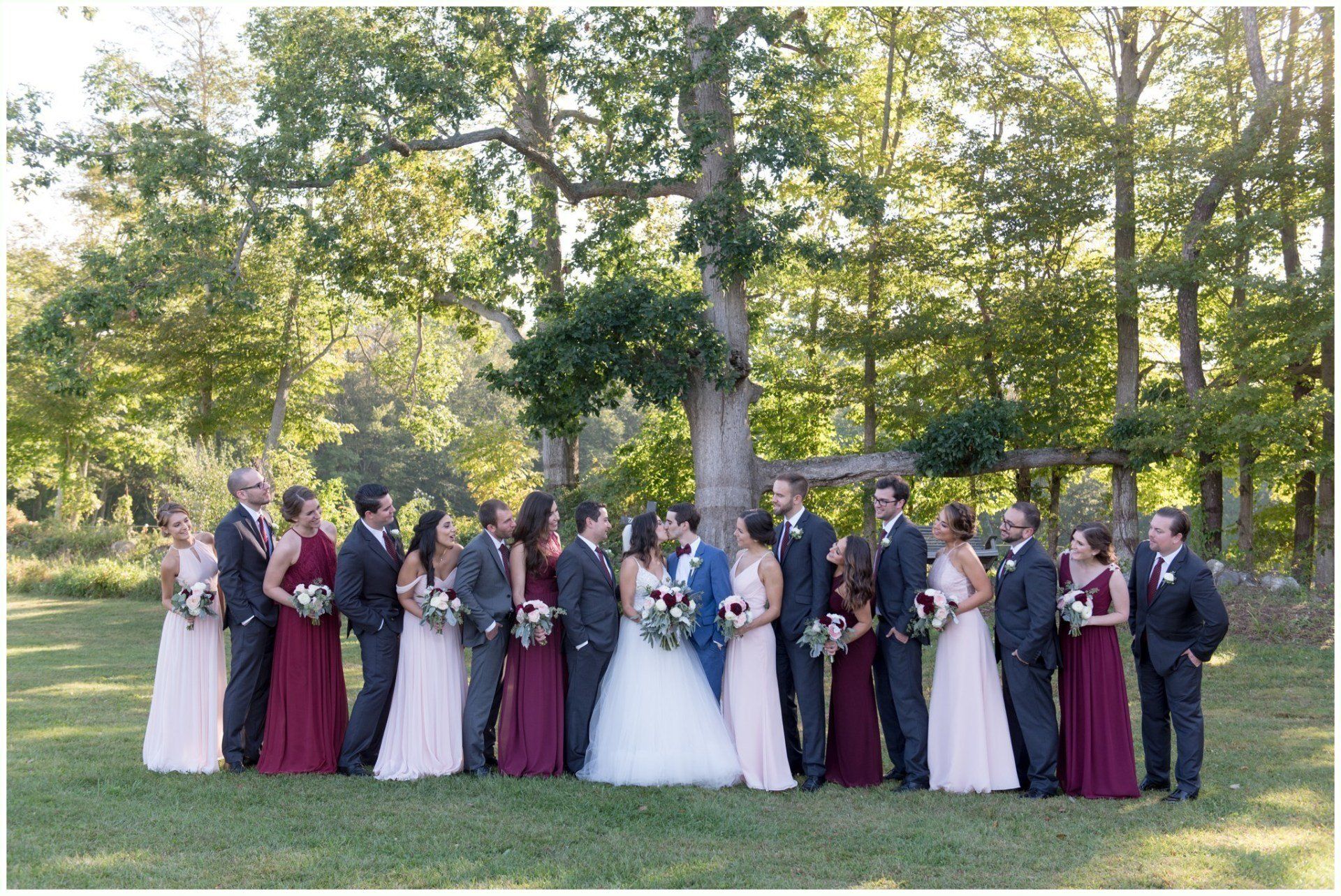 Wedding party posing outdoors in a wooded park, with bride in white and attendants in burgundy and dark suits