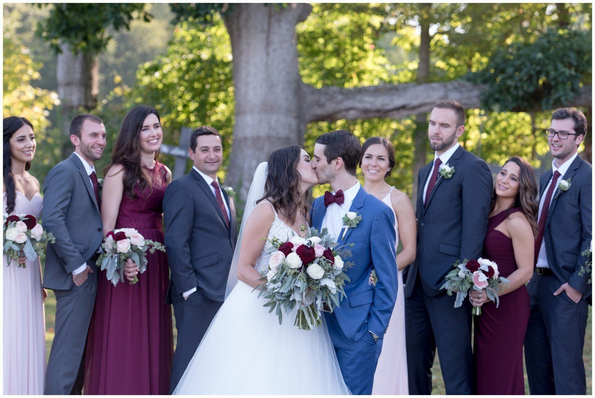 Wedding party posing outdoors, bride and groom centered, attendants in formal navy and burgundy outfits holding bouquets.