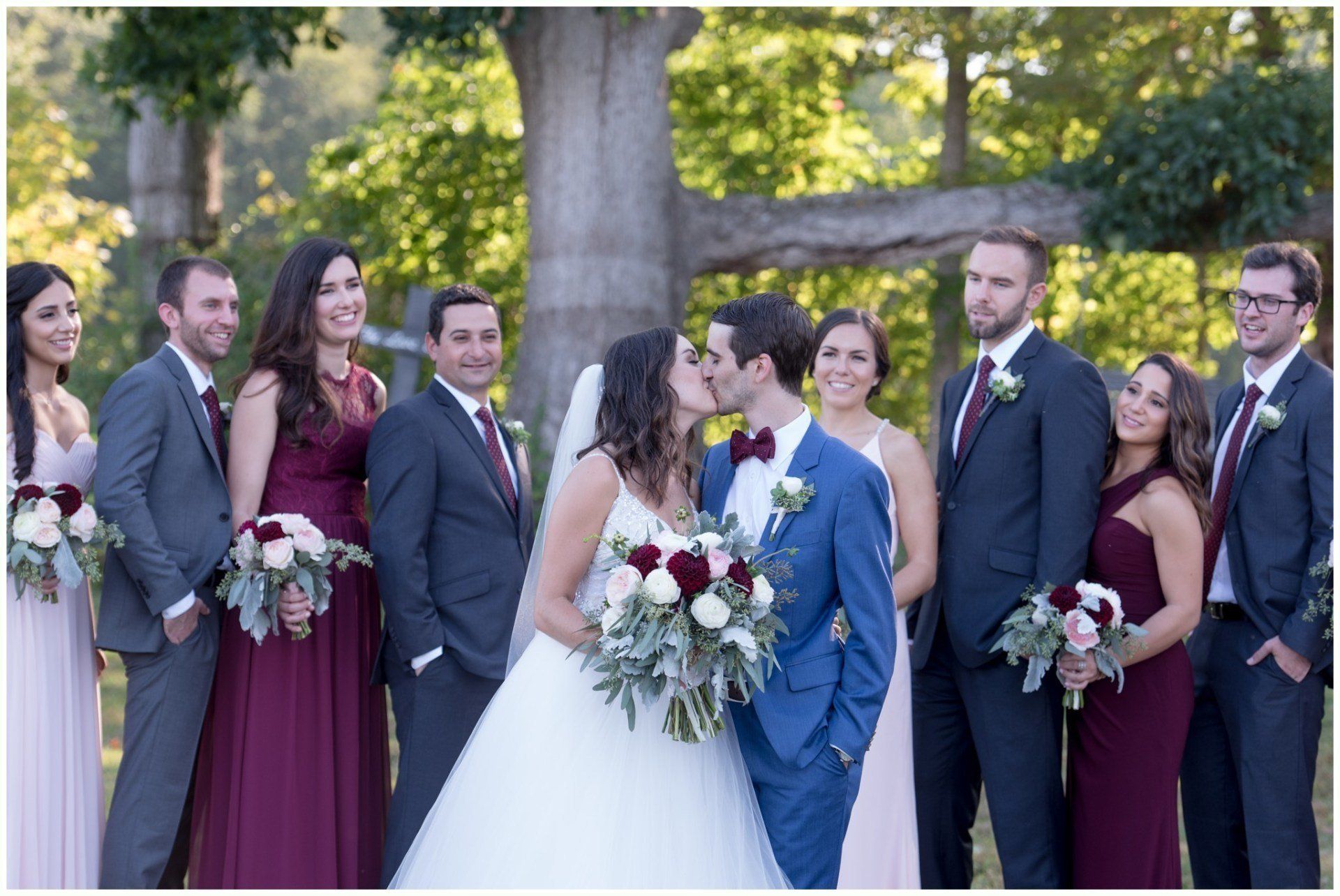 Wedding party posing outdoors among trees, with the bride in white and groom in blue at center.