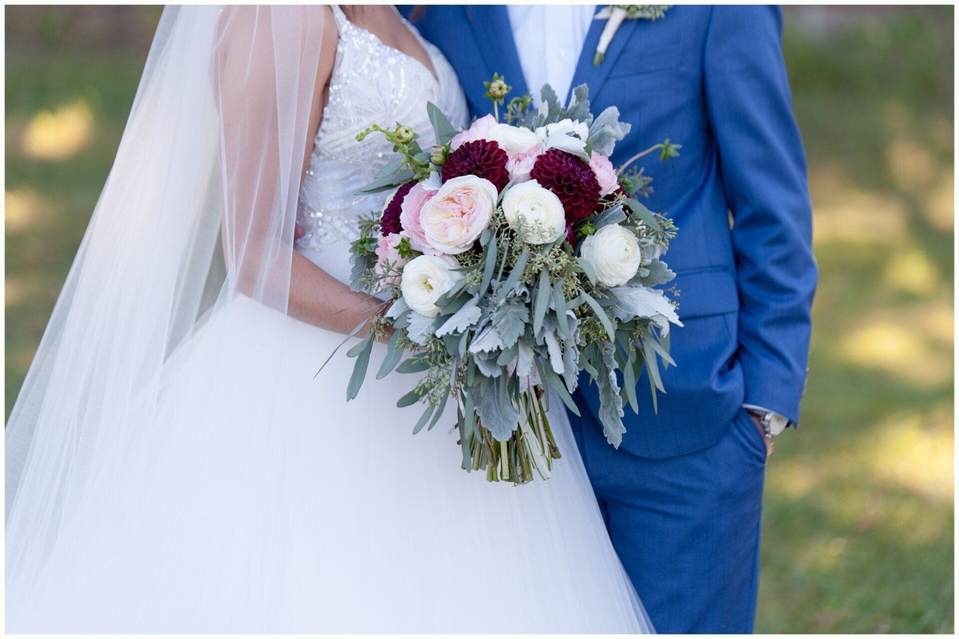Bride and groom in blue suit holding a white and burgundy wedding bouquet outdoors