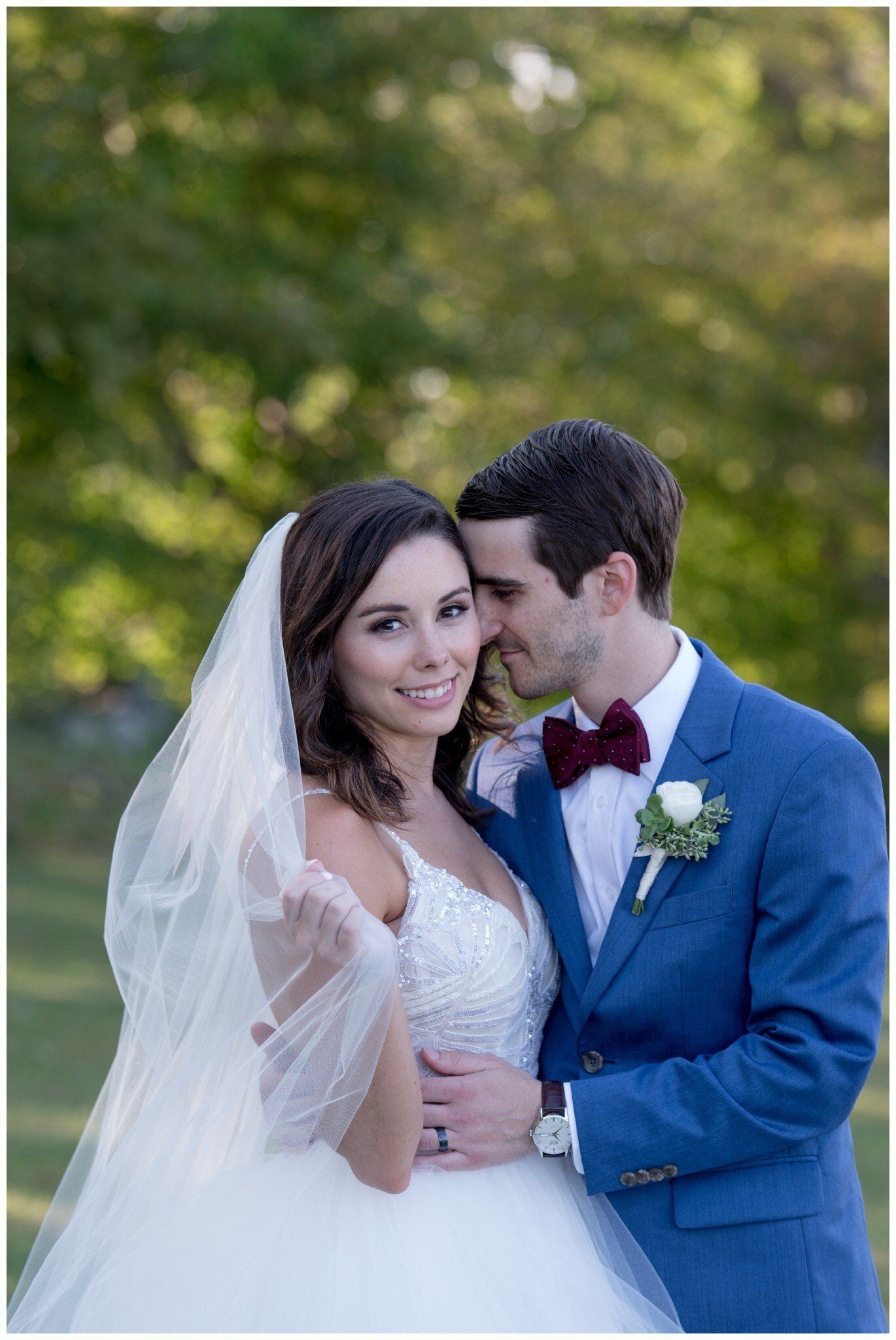 Bride and groom smiling outdoors, the groom in a blue suit and red bow tie, the bride in a white gown and veil.
