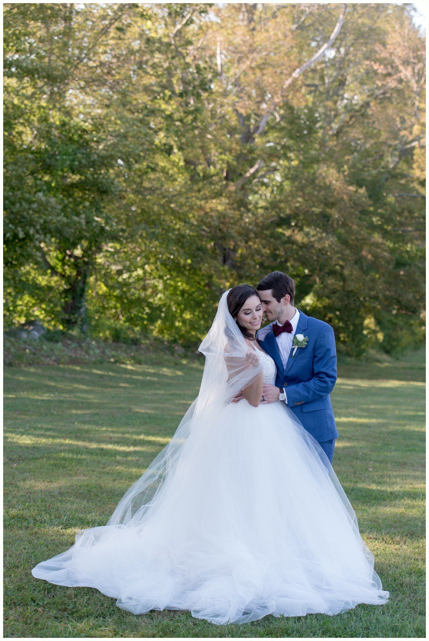 Bride and groom embracing outdoors in a grassy park, bride in white gown and veil, groom in blue suit.