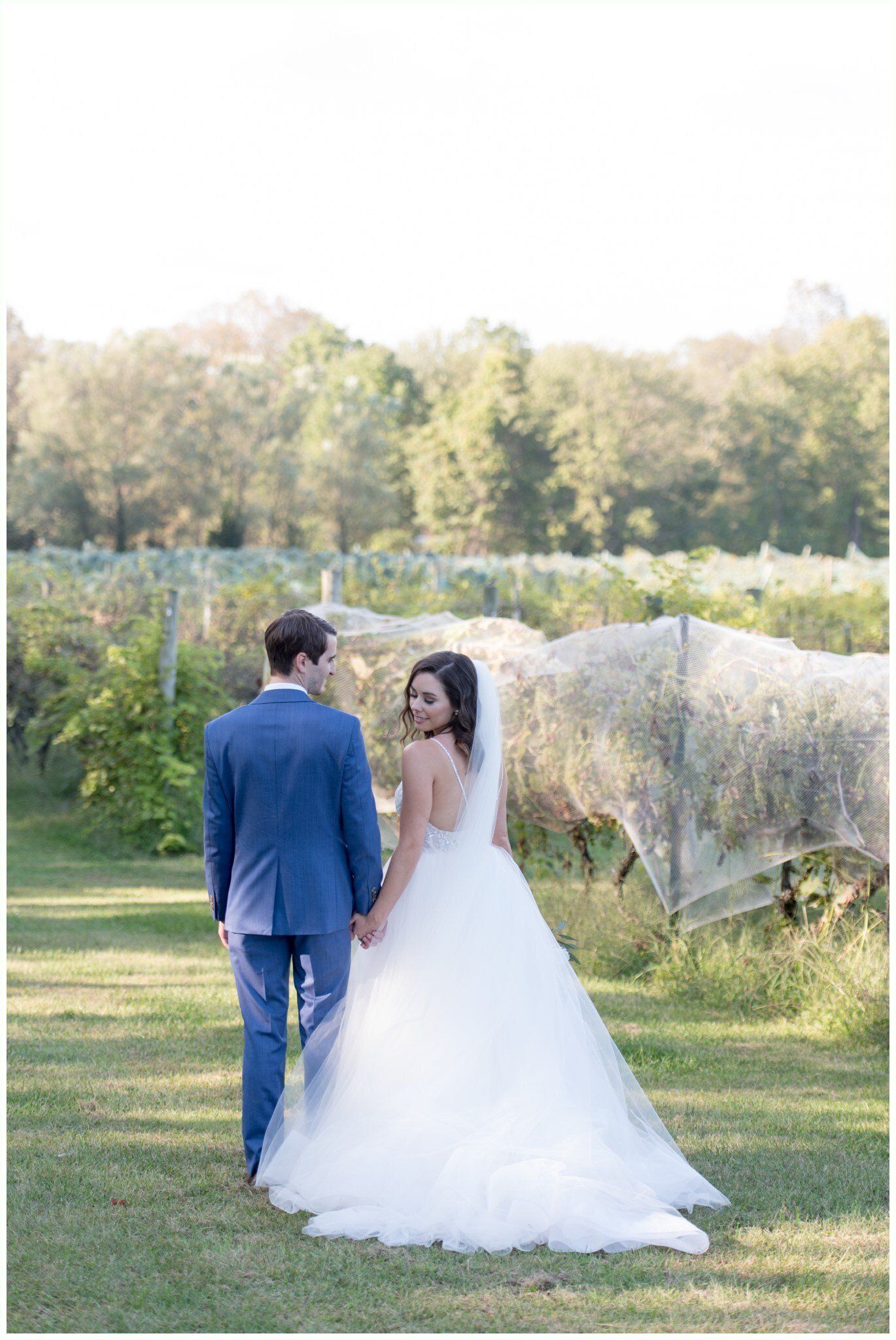 Bride and groom walking hand in hand on a grassy field beside a pond, with trees in the background