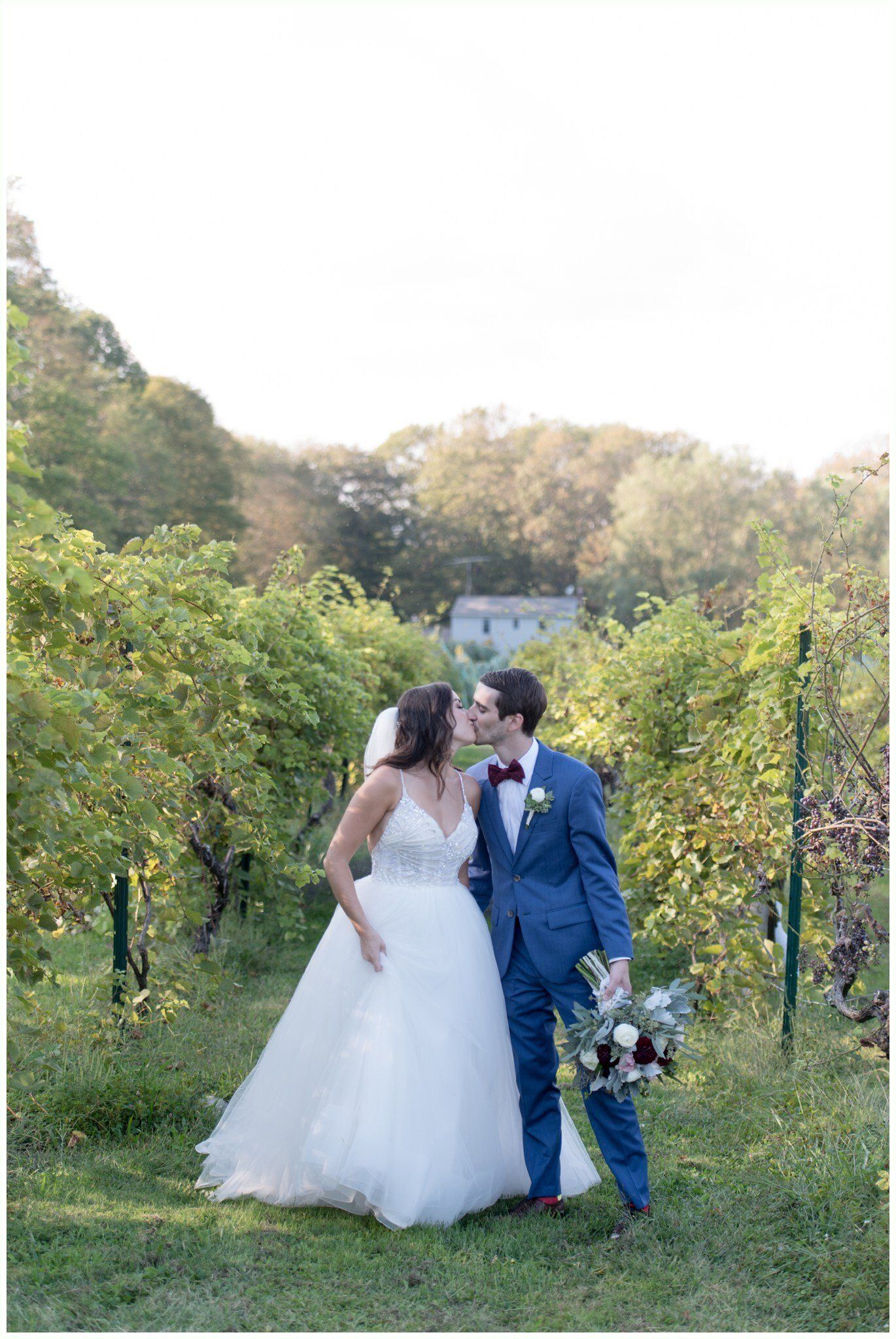 Wedding couple kissing in a vineyard, bride in white gown and groom in blue suit holding bouquet