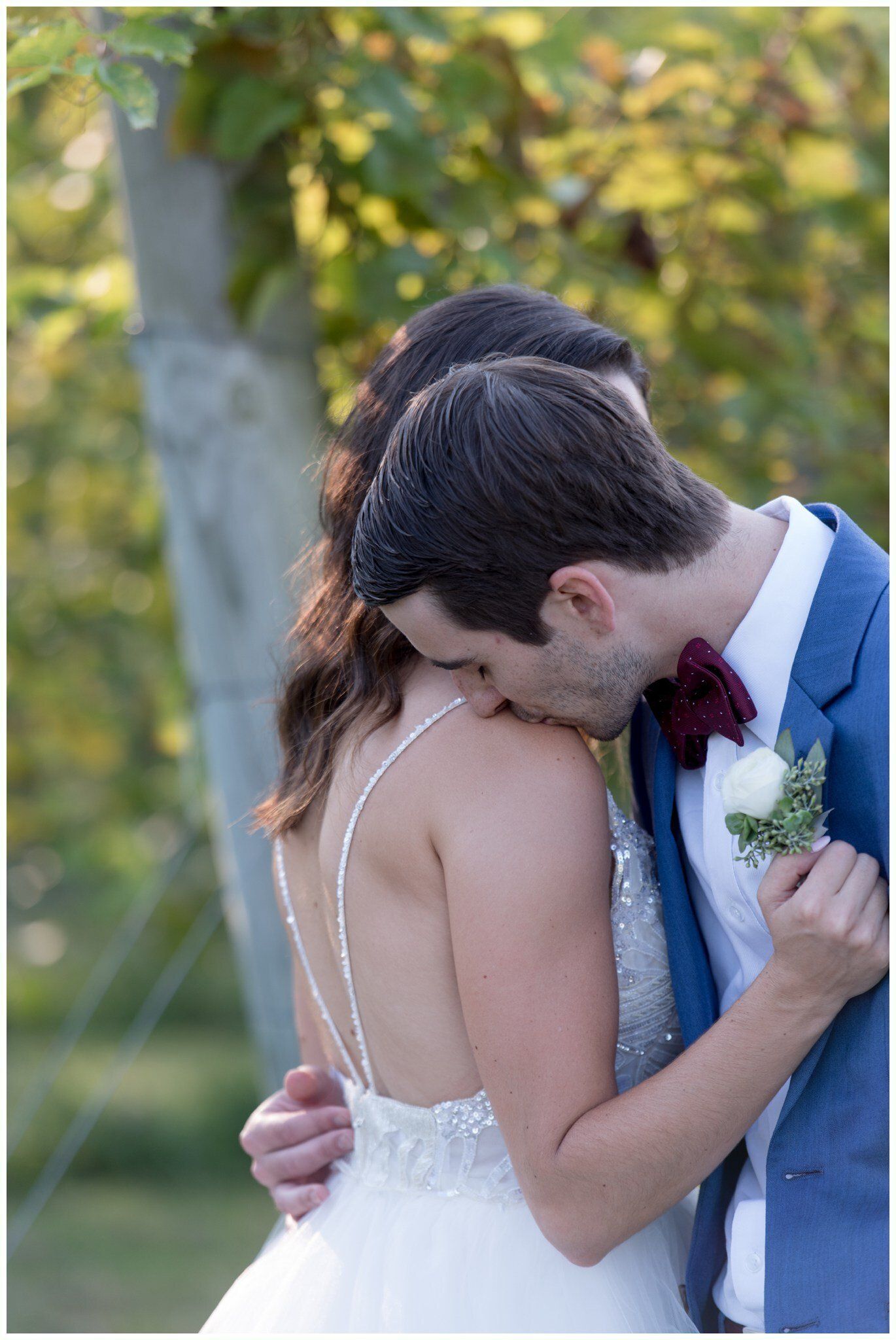Couple embracing outdoors at a wedding, groom in blue suit kissing bride in white dress on the shoulder