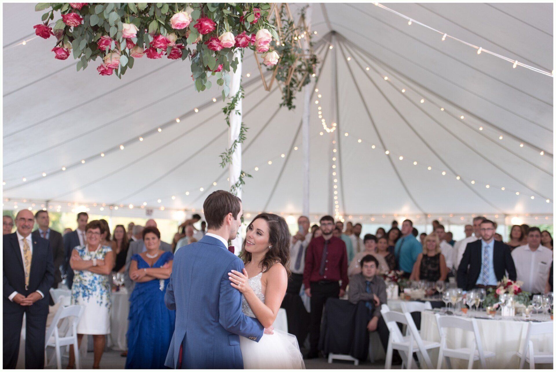 Couple dancing at a wedding reception under a white tent with string lights and floral decorations