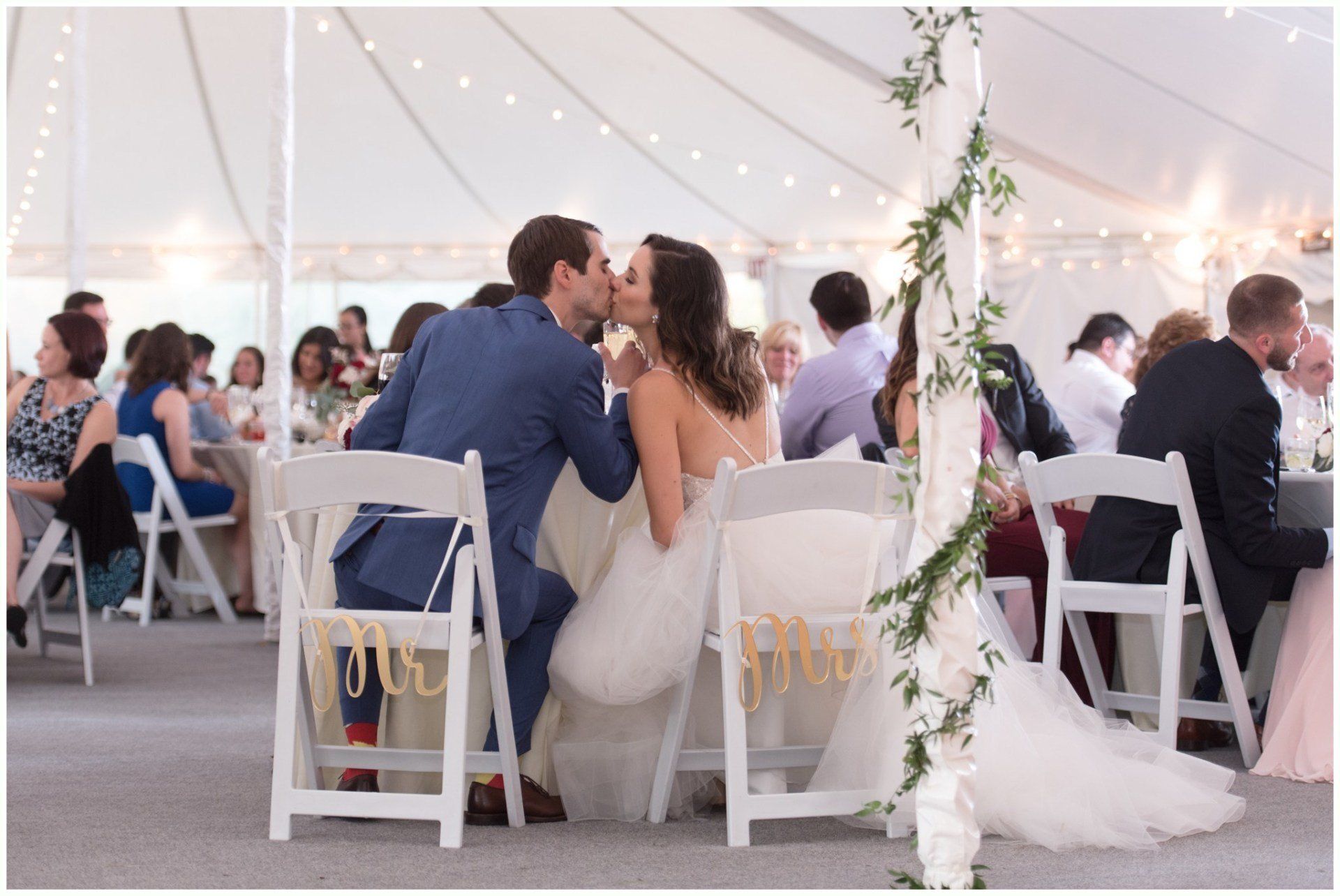 Couple kissing at a decorated outdoor wedding reception under a white tent with string lights