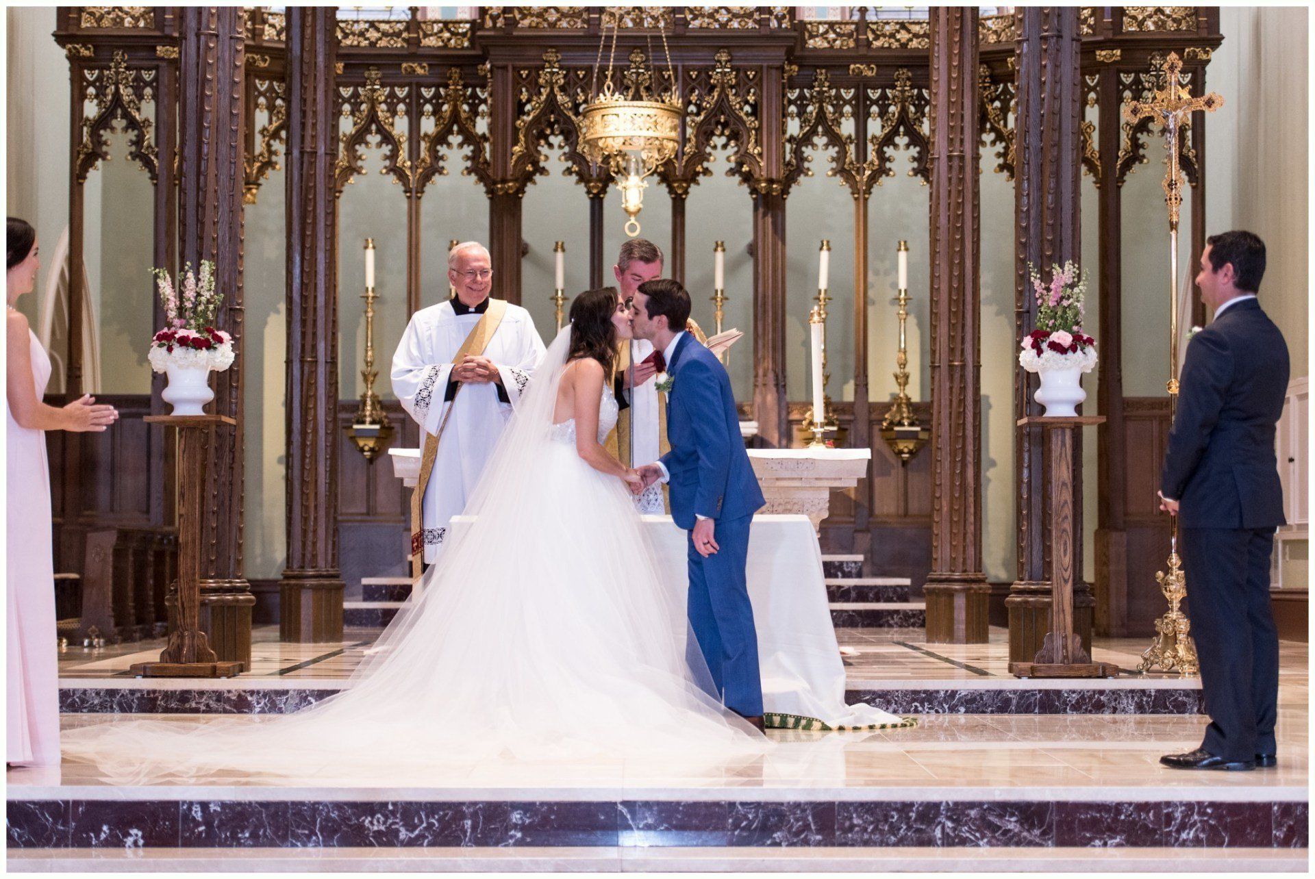 Bride and groom kiss at the altar during a church wedding ceremony, with officiant and guests nearby