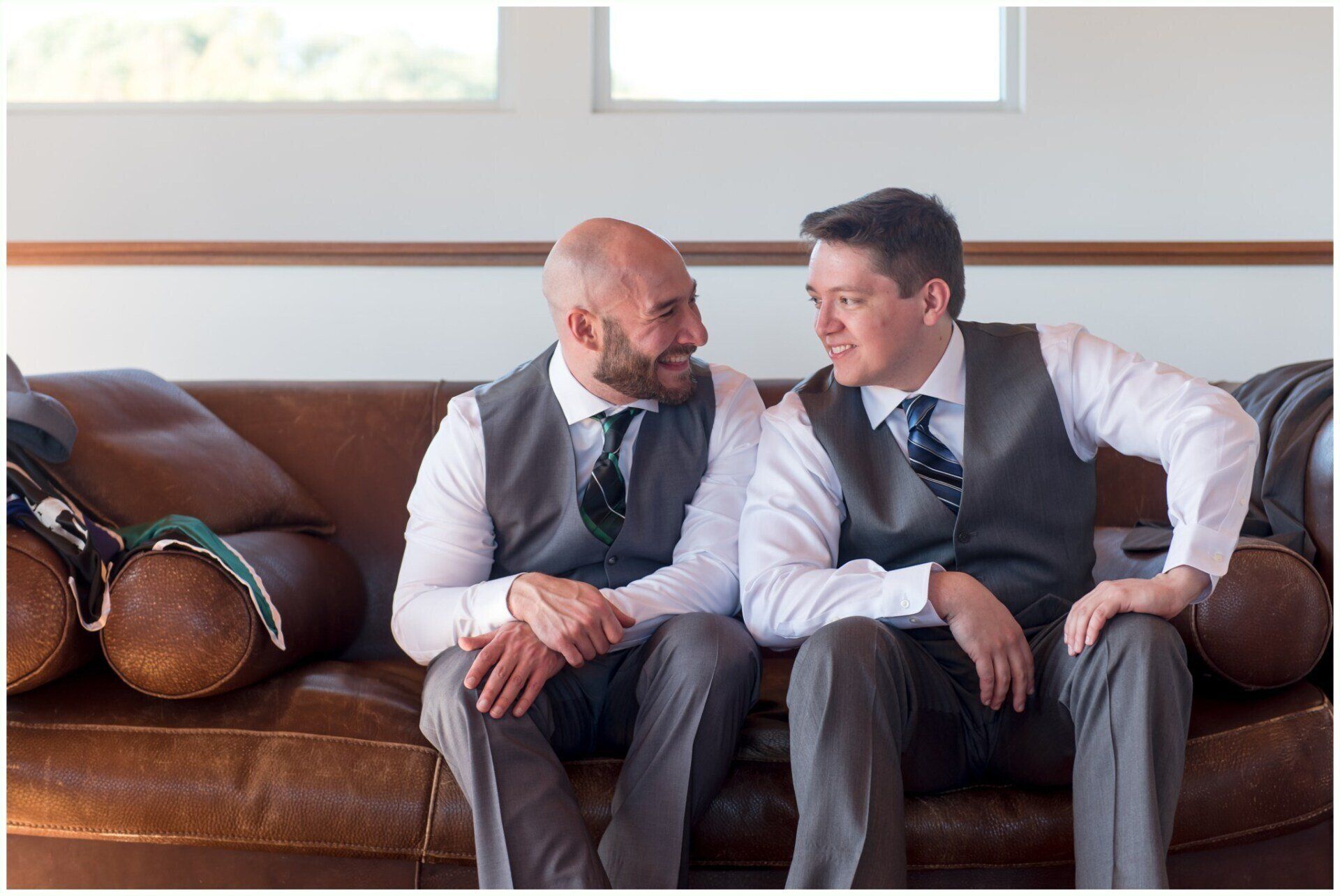 Two men in suits sit on a couch talking, with a bag on the armrest beside them.