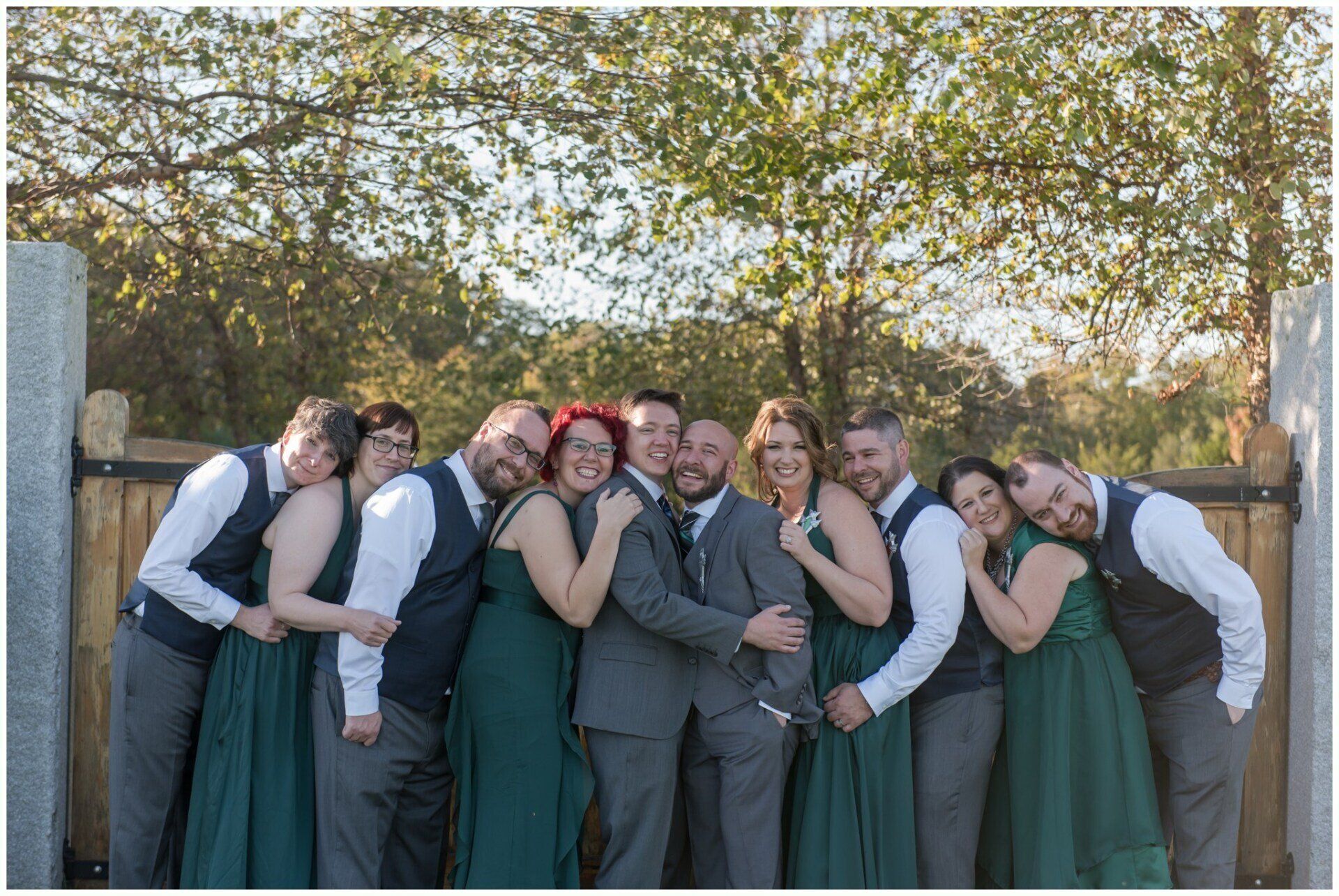 Group of people in formal attire posing together outdoors, smiling and hugging in front of trees.