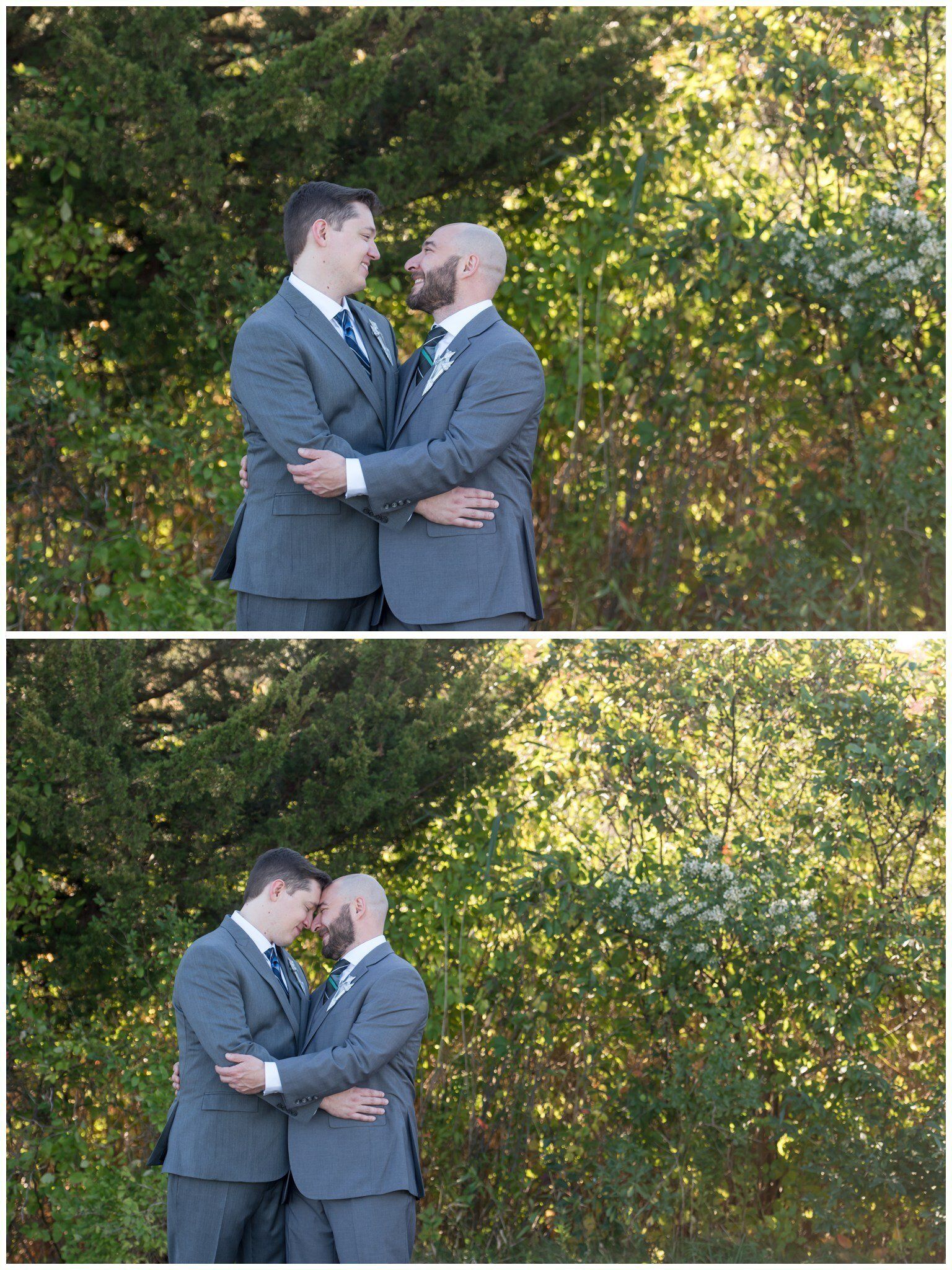Two grooms in gray suits embrace and kiss outdoors in front of sunlit greenery.