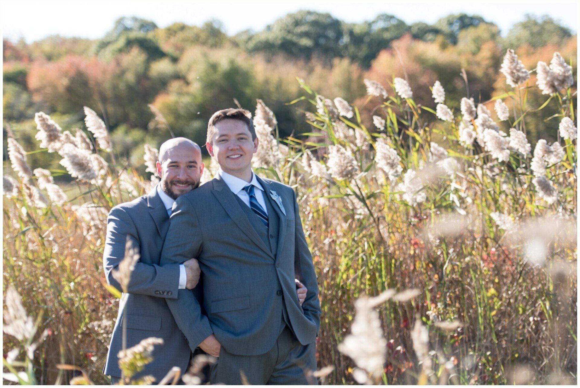 Two men in suits smiling in a sunlit field with tall grass and autumn trees