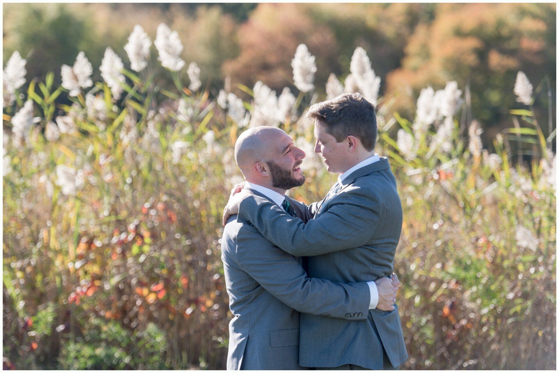 Two men in blue suits embrace outdoors amid tall white flowers and greenery.