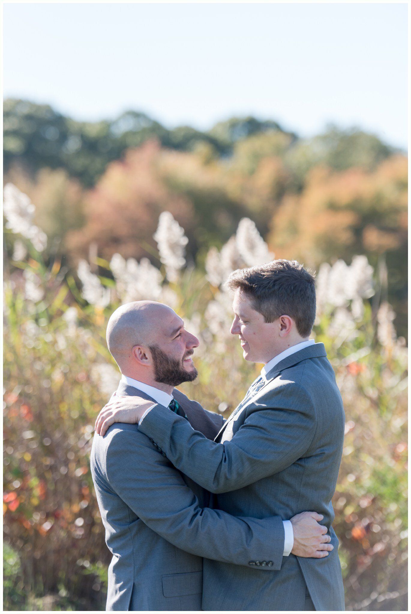 Two men in suits embracing outdoors in a sunlit field with tall grasses and autumn trees
