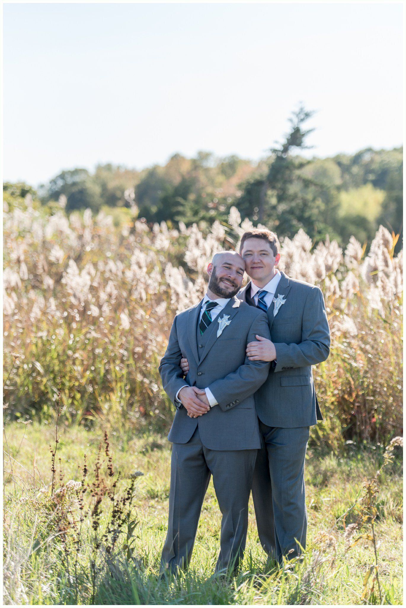 Two people in gray suits posing together in a sunny field with tall grass and trees.