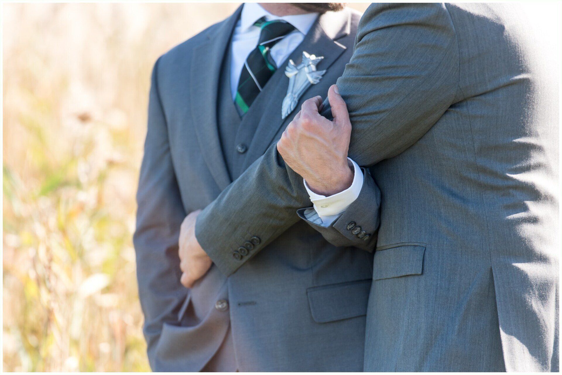 Two men in gray suits standing arm in arm outdoors, one holding a bouquet with a striped tie visible.