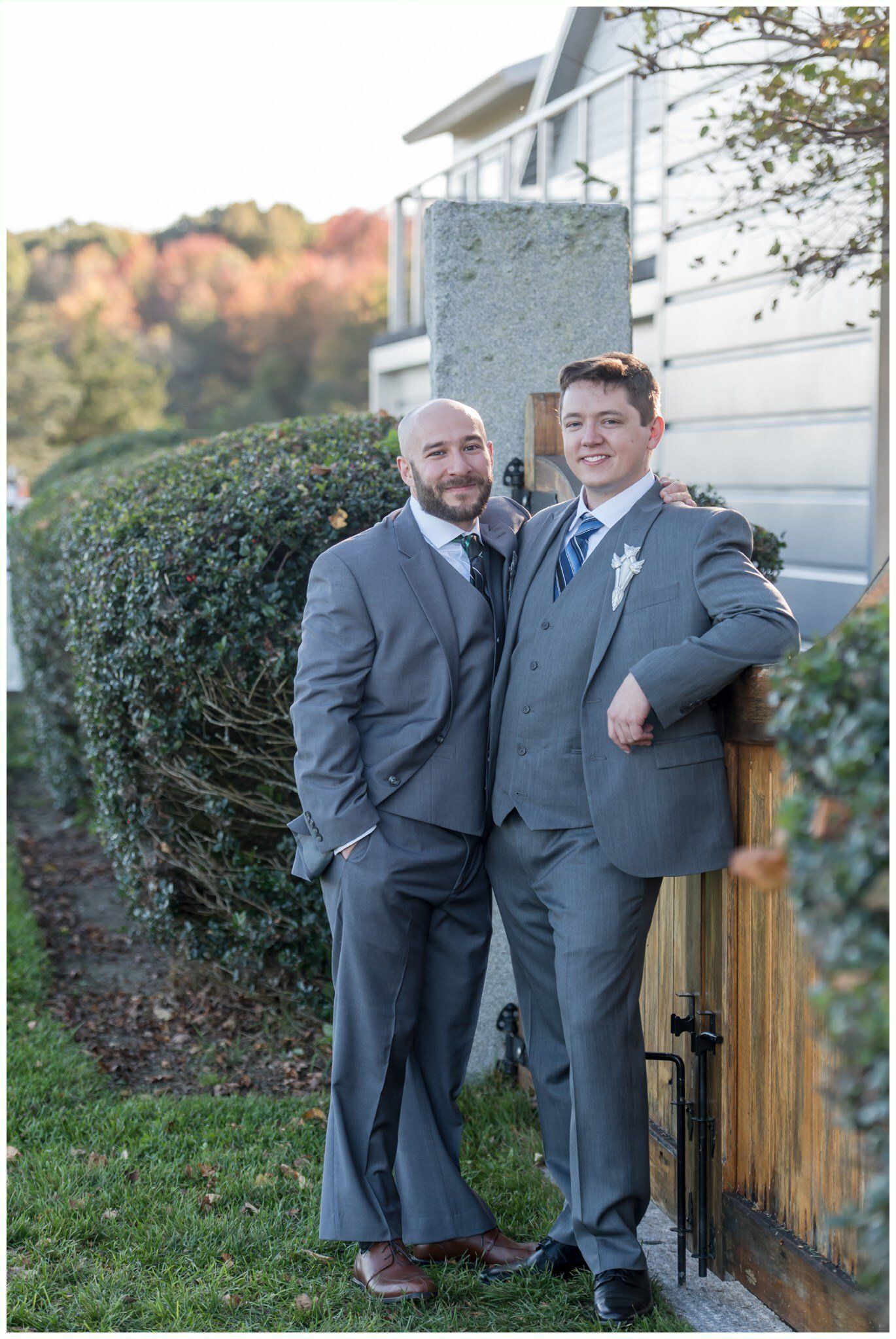 Two men in gray suits standing together outdoors beside a hedge and house