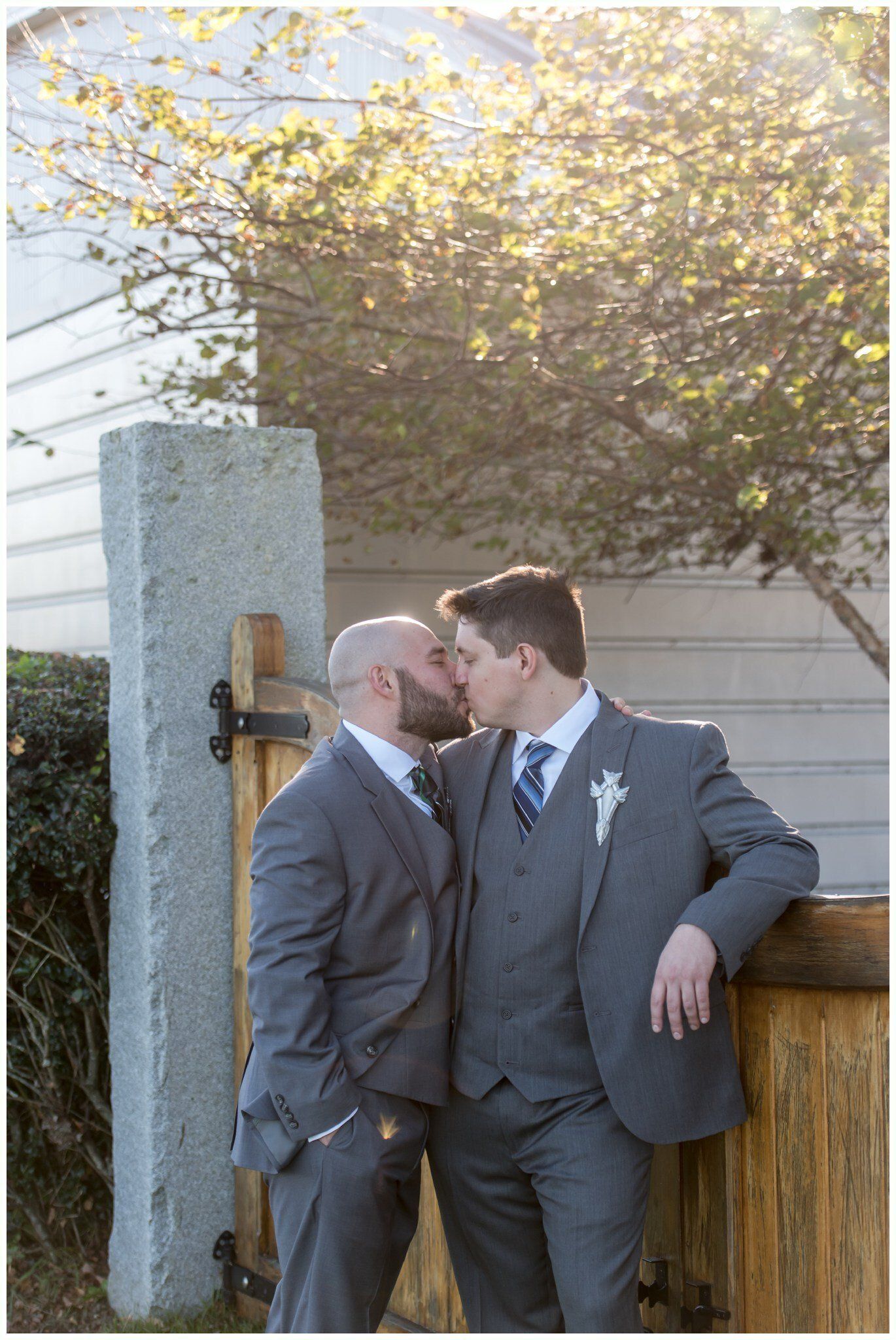 Two men in gray suits kissing outdoors beside a wooden fence and house
