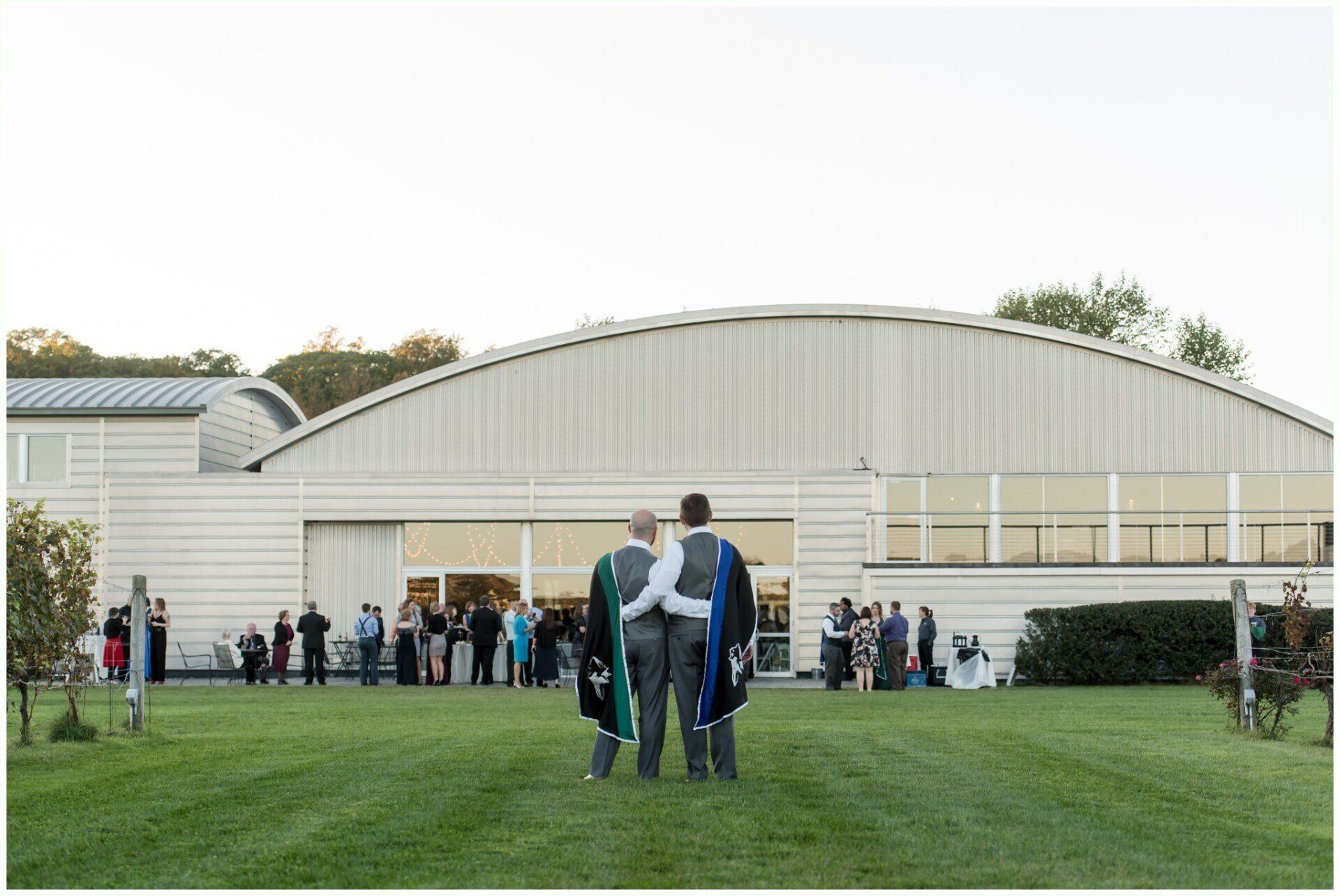 People gathered outside a large white building on a grassy lawn, with three people in dark robes in the foreground.