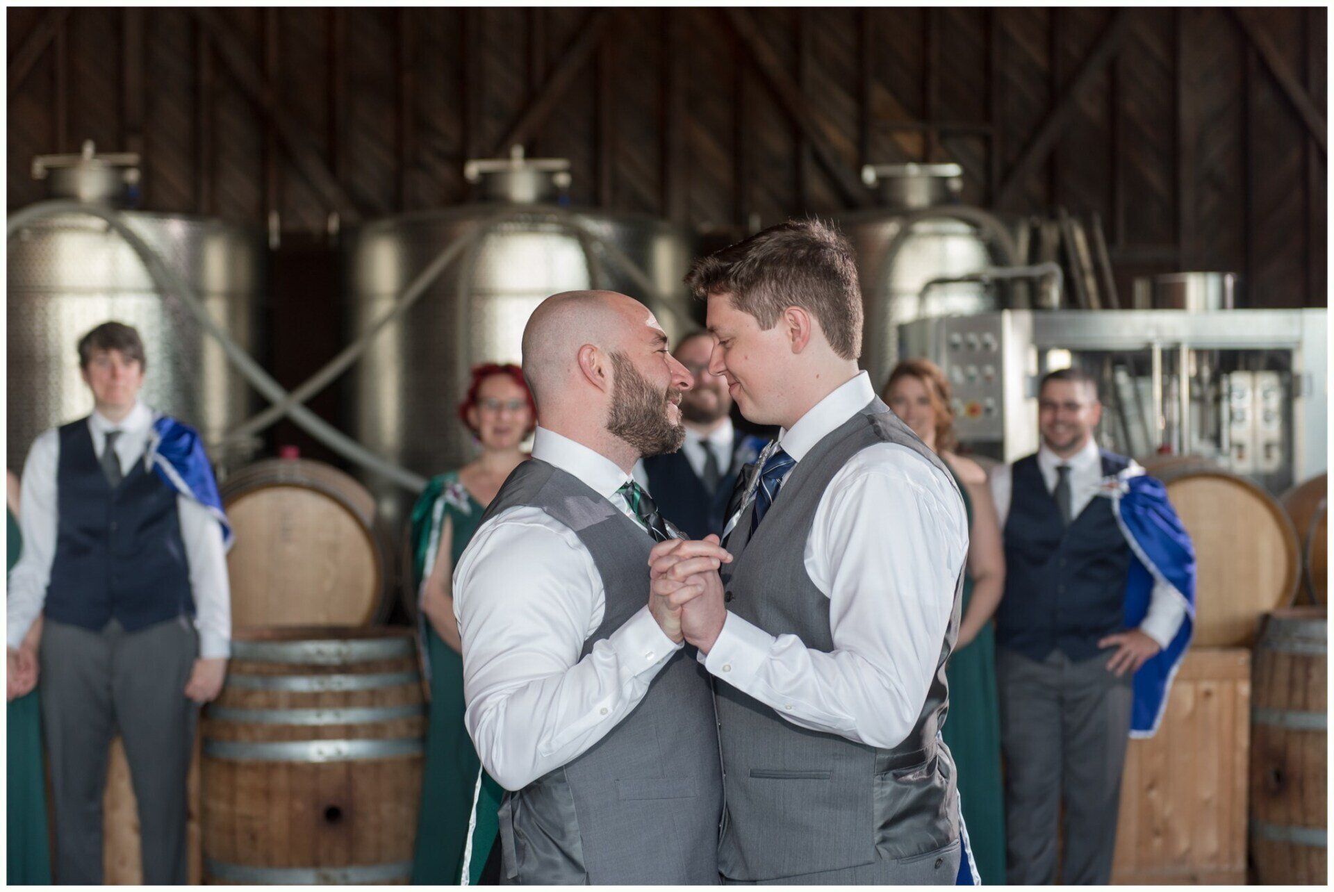 Two men in vests dancing closely in a winery, with three onlookers in the background
