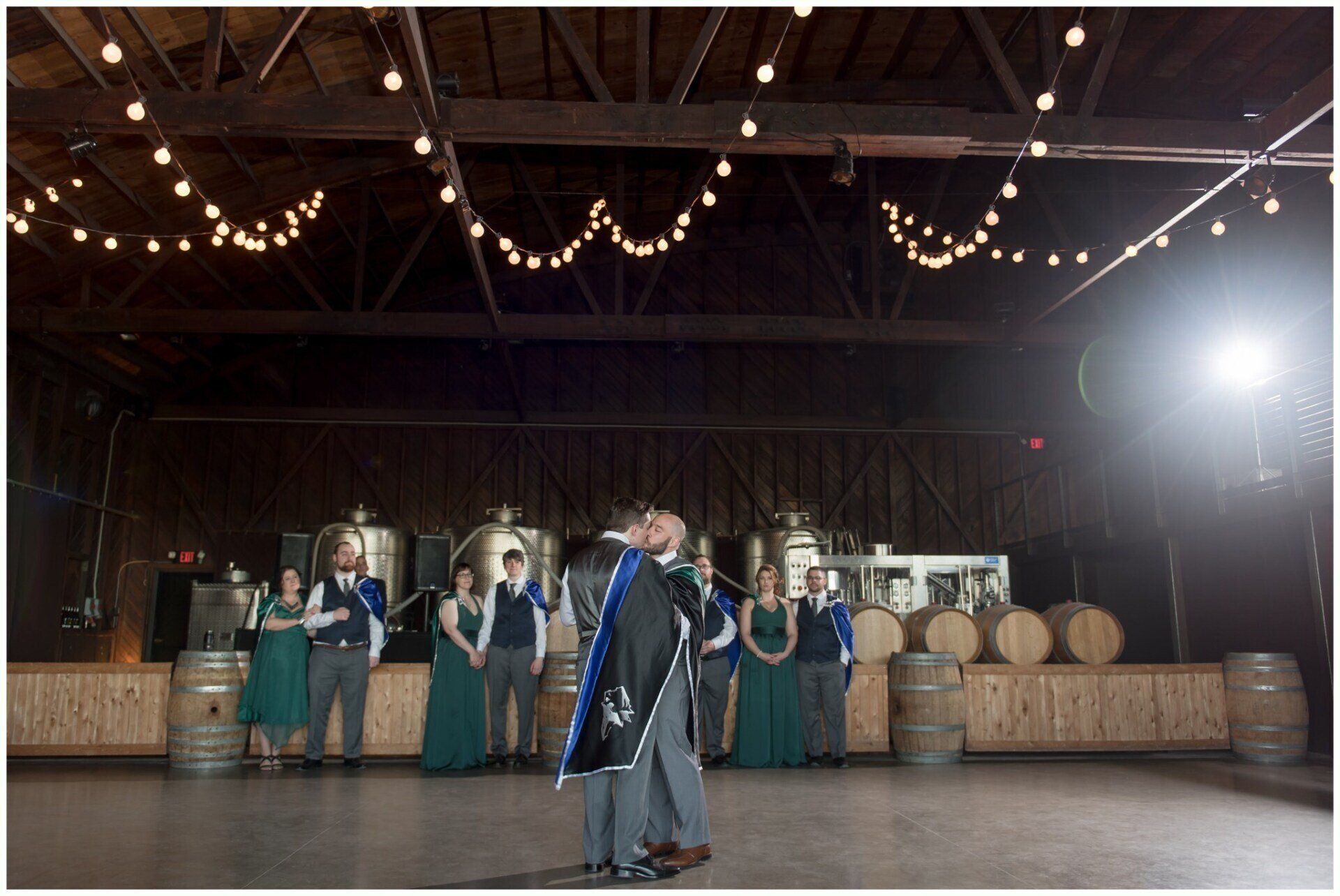People in teal robes perform on a barn-like stage with string lights and barrels in the background.