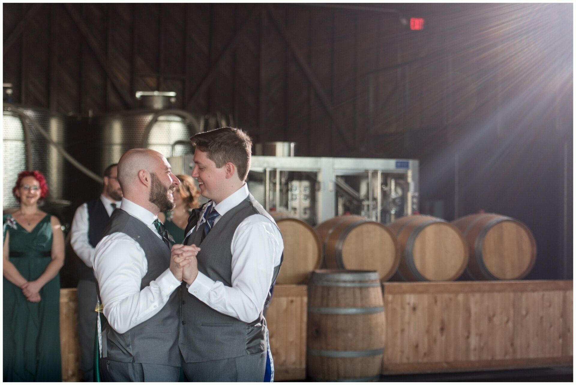 Two people in formal attire dance in a rustic winery with barrels and warm backlighting.