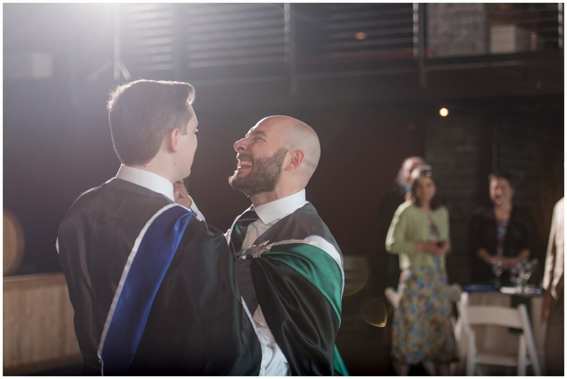 Two graduates in caps and gowns face each other at a ceremony, with guests blurred in the background.