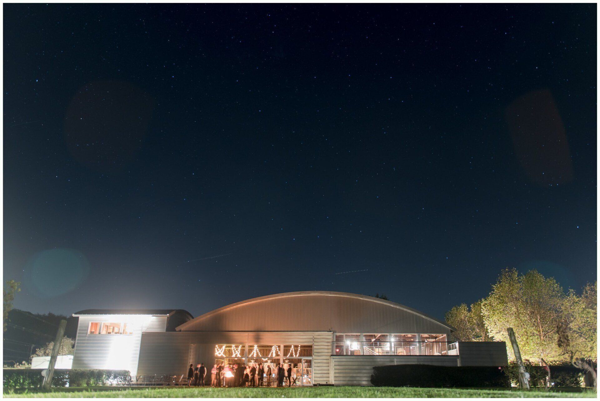 Nighttime view of a lit modern building with a curved roof and people gathered at the entrance.