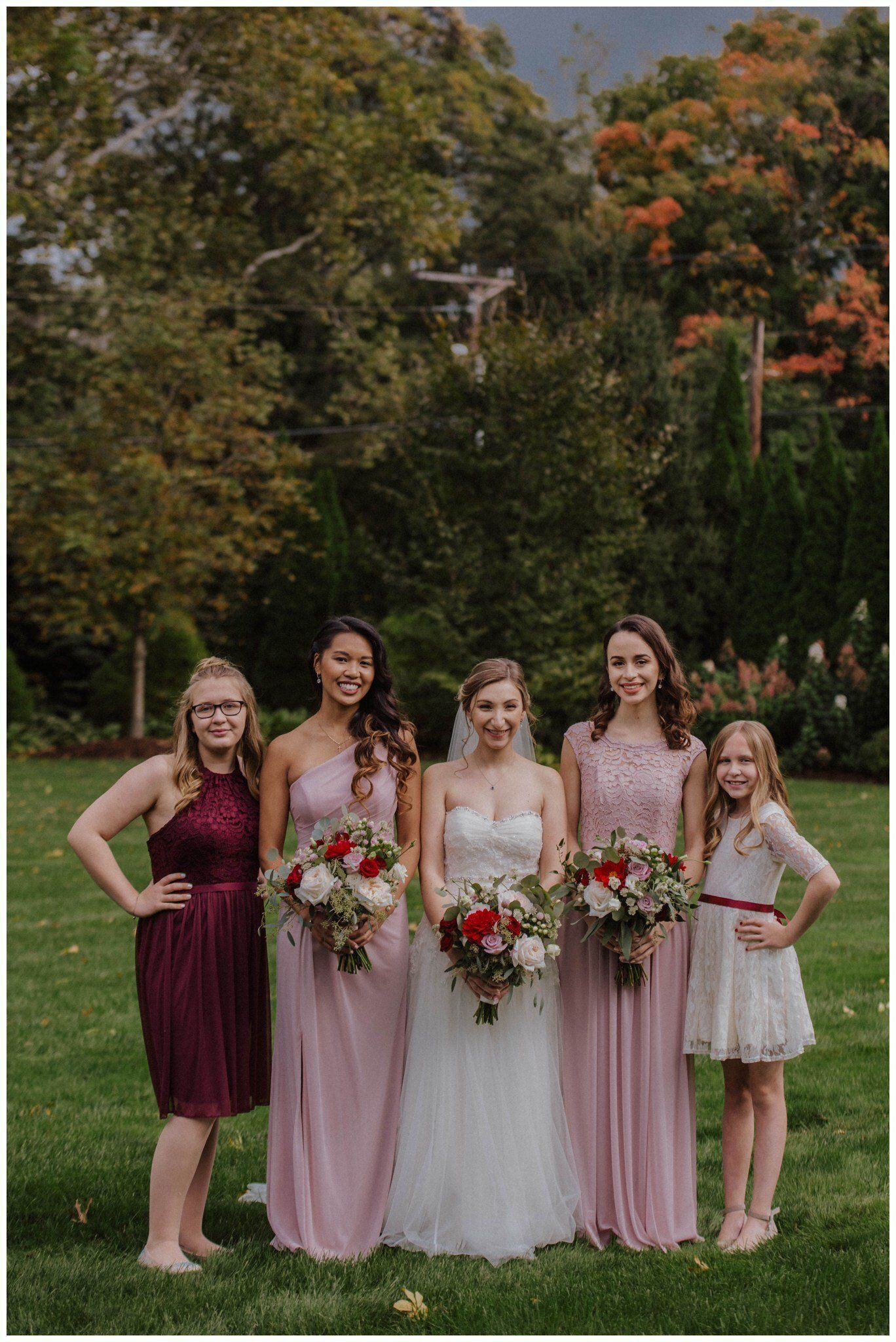 Five women in dresses holding bouquets pose on a lawn in a garden.