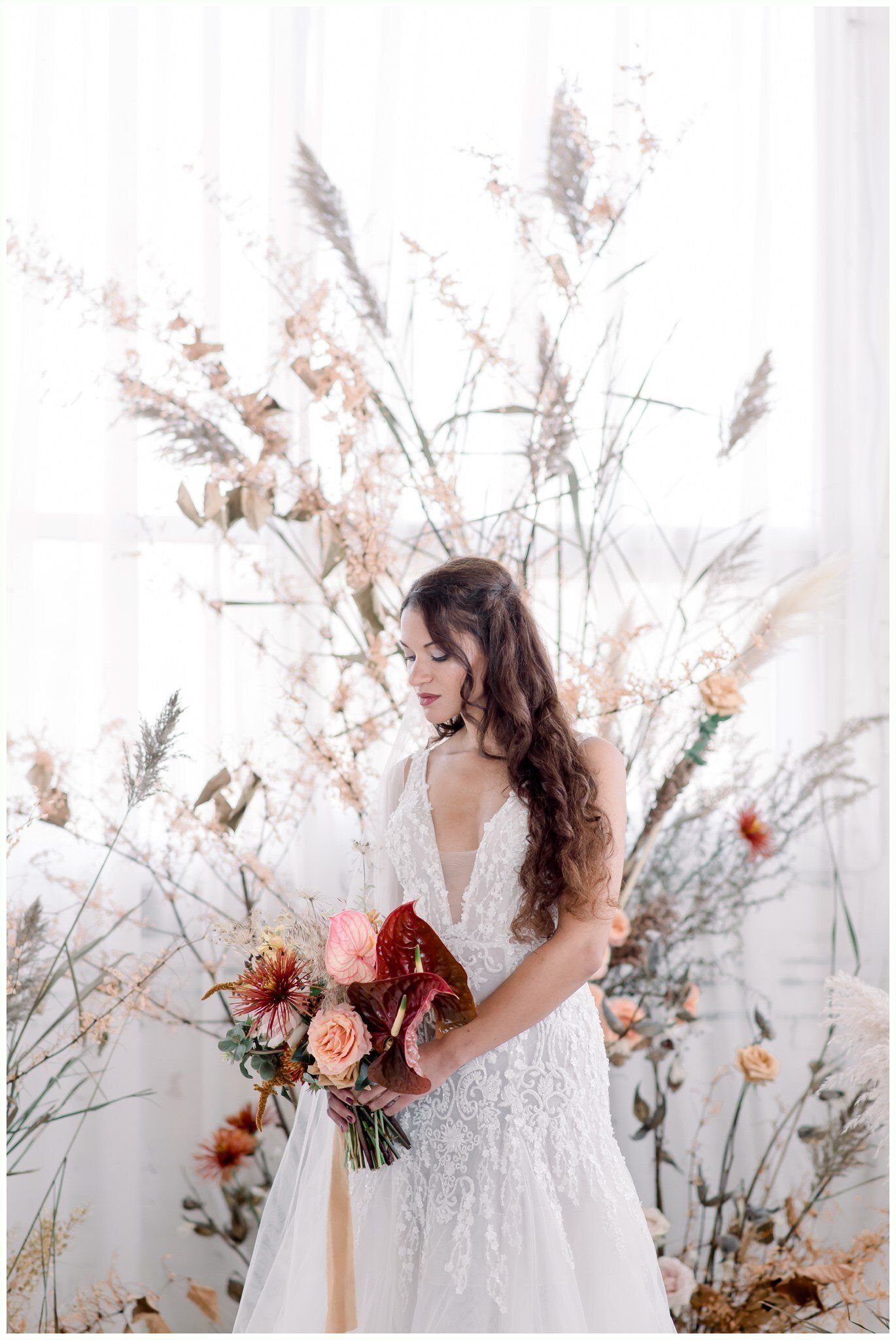 Bride in a white gown holding a bouquet, standing before a floral backdrop in soft light