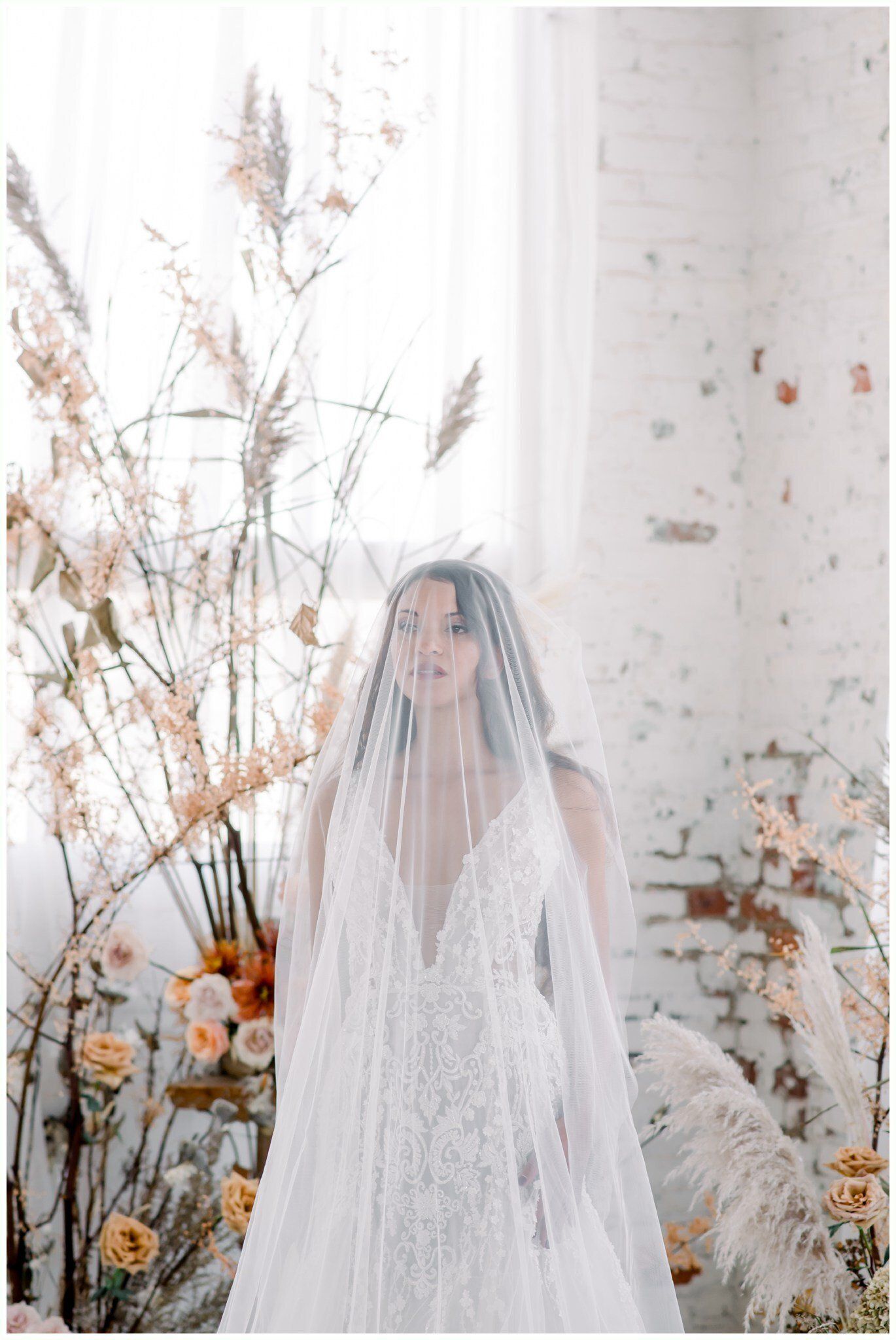 Bride in a white lace gown and veil, standing among dried flowers against a pale backdrop