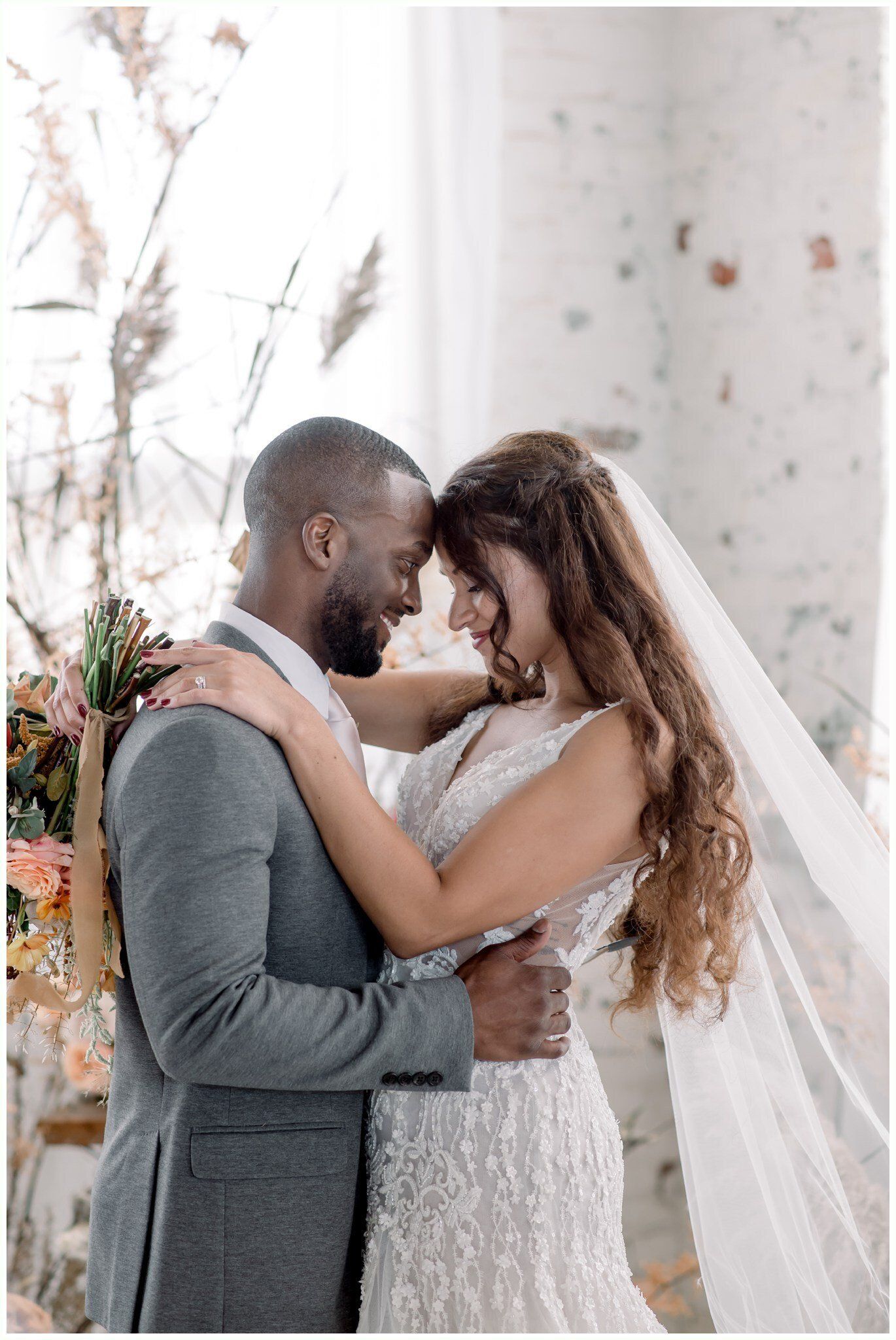 Couple embracing in wedding attire beside a white column, with a sheer veil draped behind them.