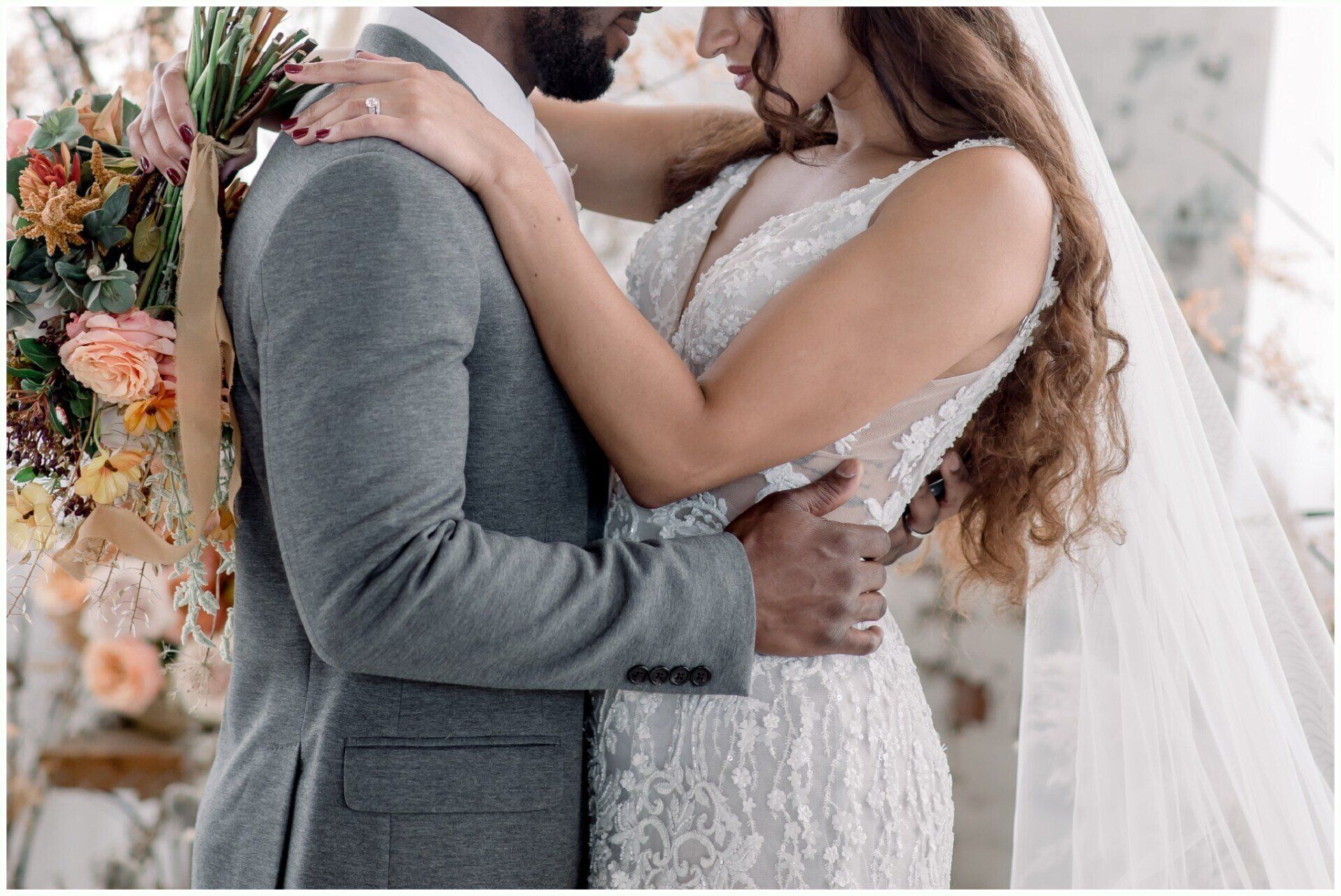 Wedding couple embracing, with bride in white lace dress and groom in gray suit, bouquet behind them.