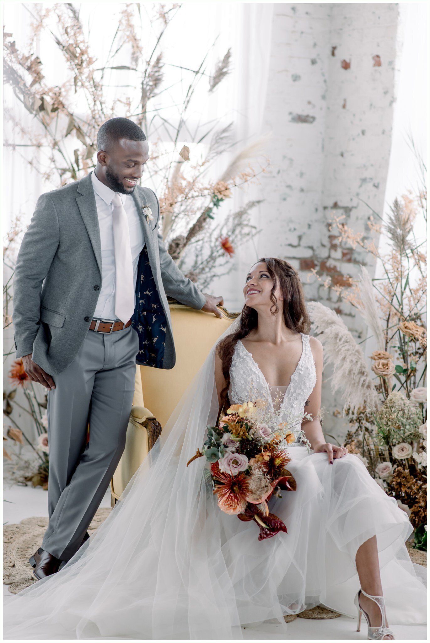 Couple in wedding attire by floral drapes, groom standing and bride seated holding a bouquet.
