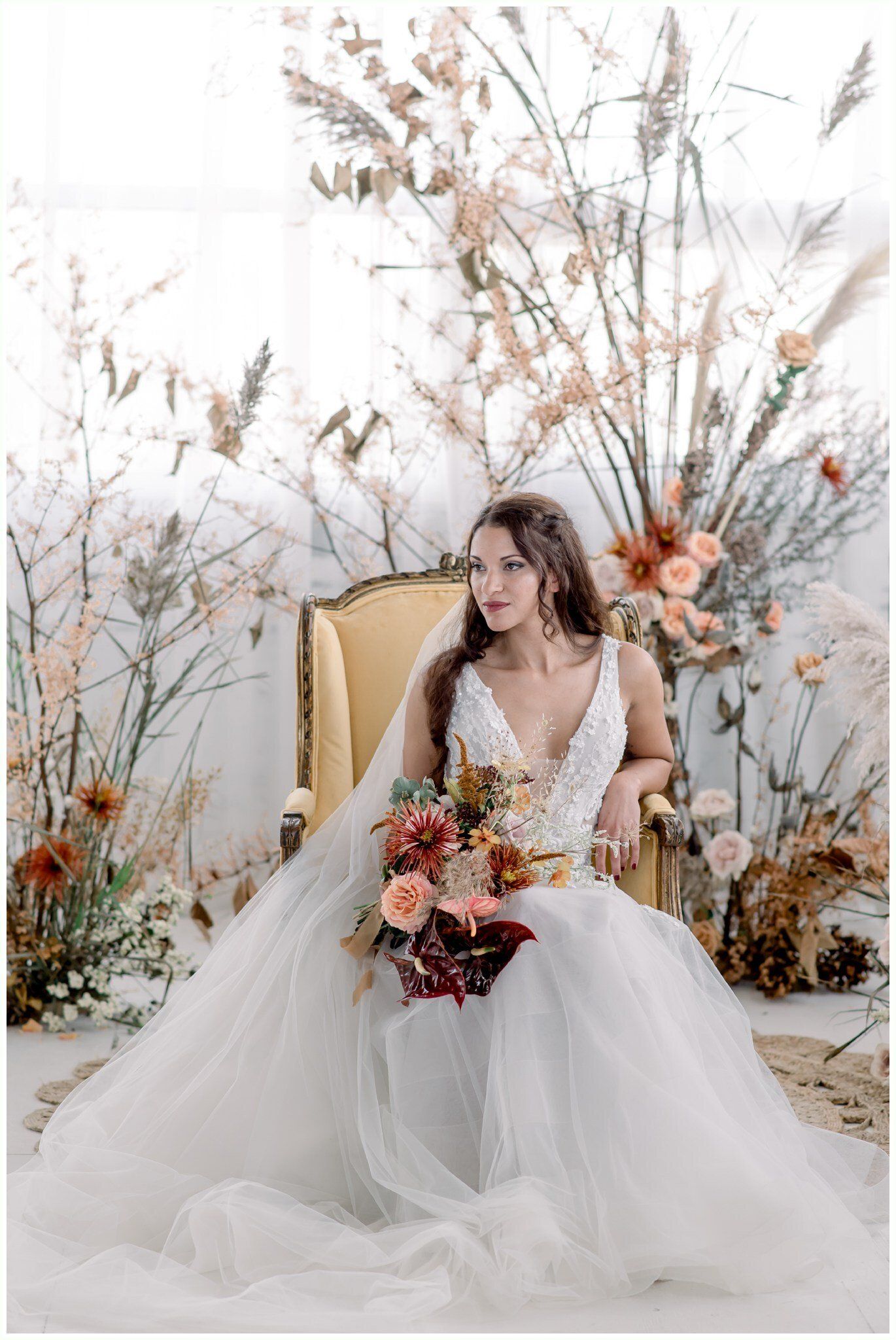 Bride in white gown seated among dried floral arrangements, holding a bouquet in a soft, airy studio setting
