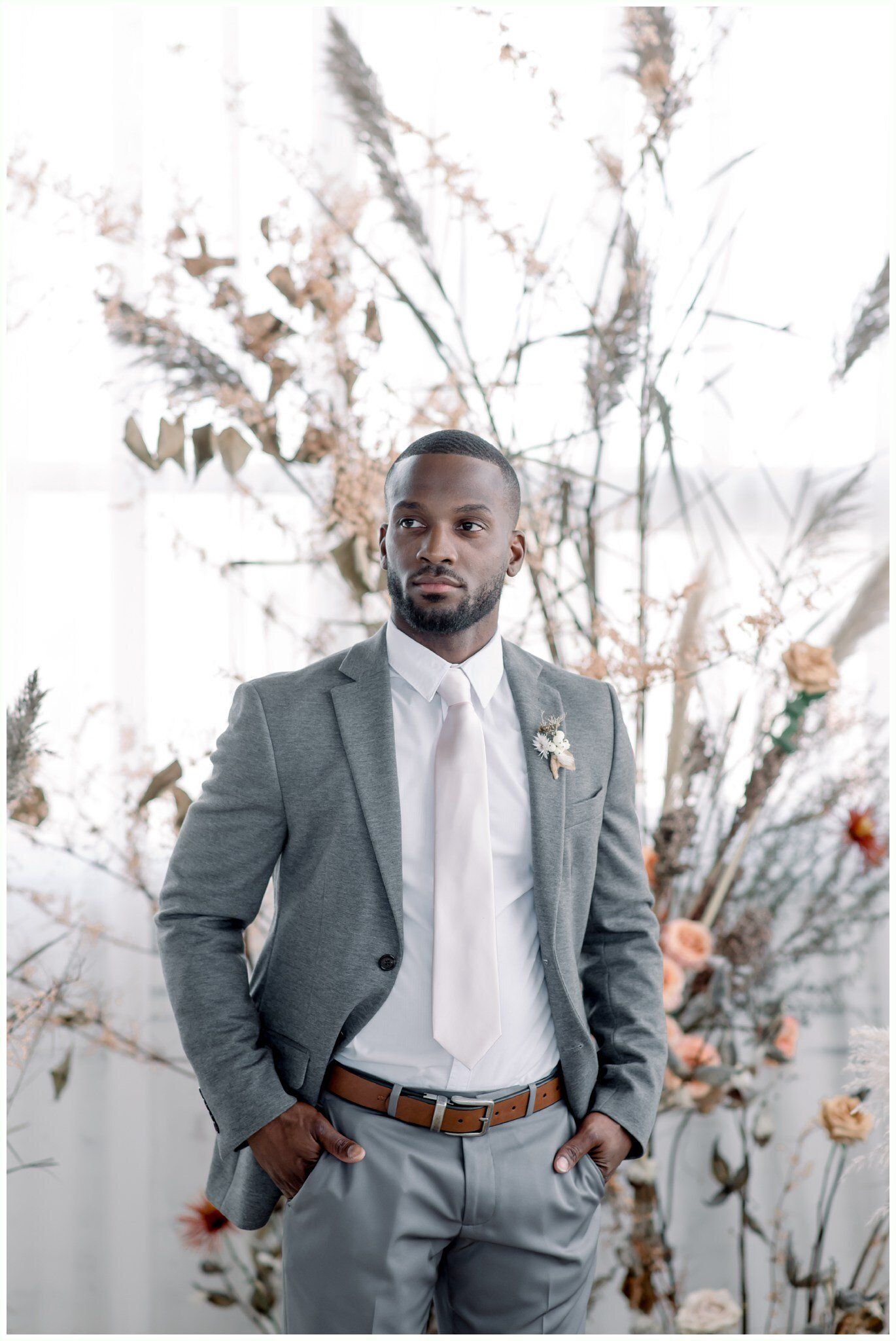 Man in a gray suit standing before white floral branches, hands in pockets, in a bright studio setting