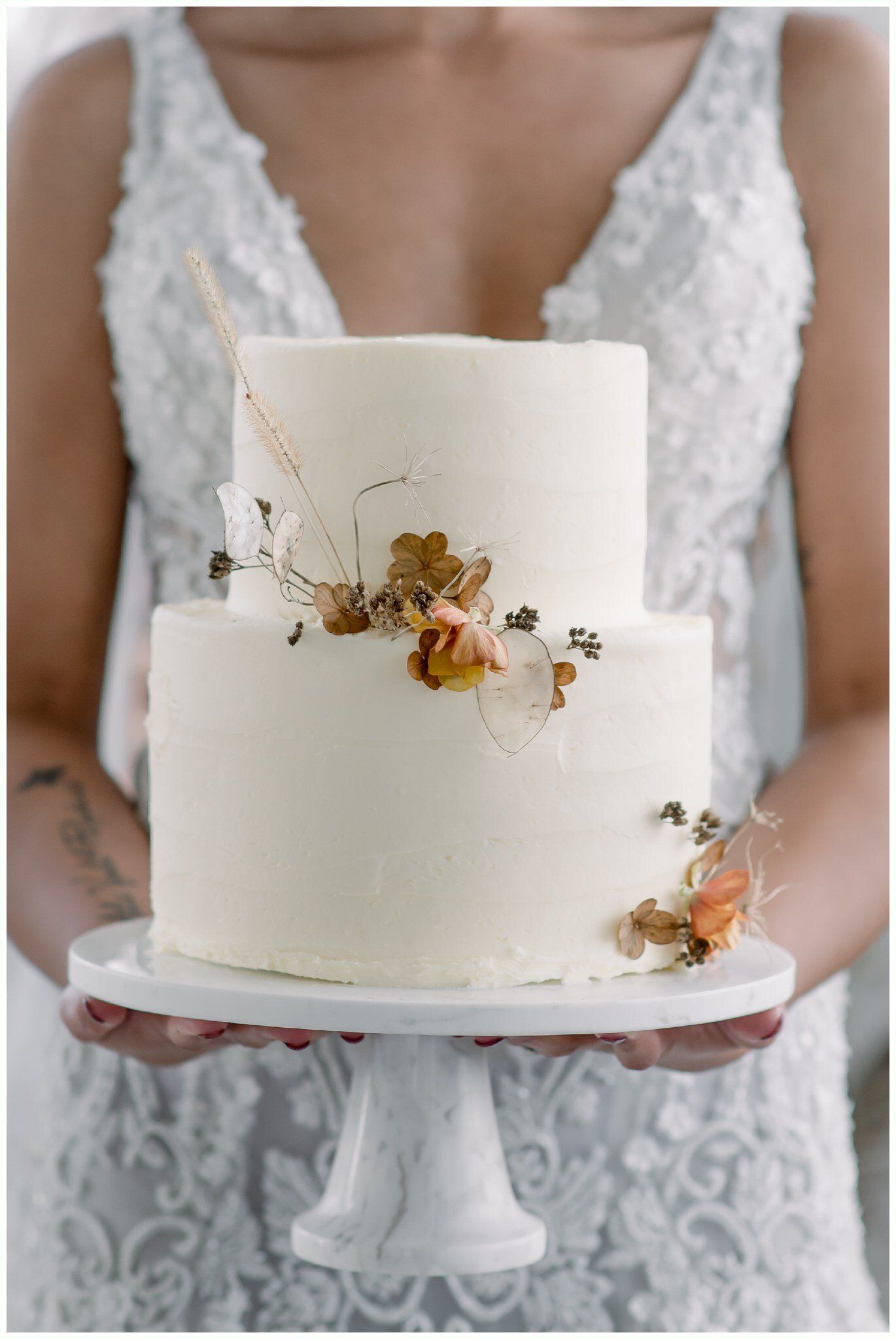 White two-tier wedding cake with dried flowers on a decorative stand, held by someone in a lace dress