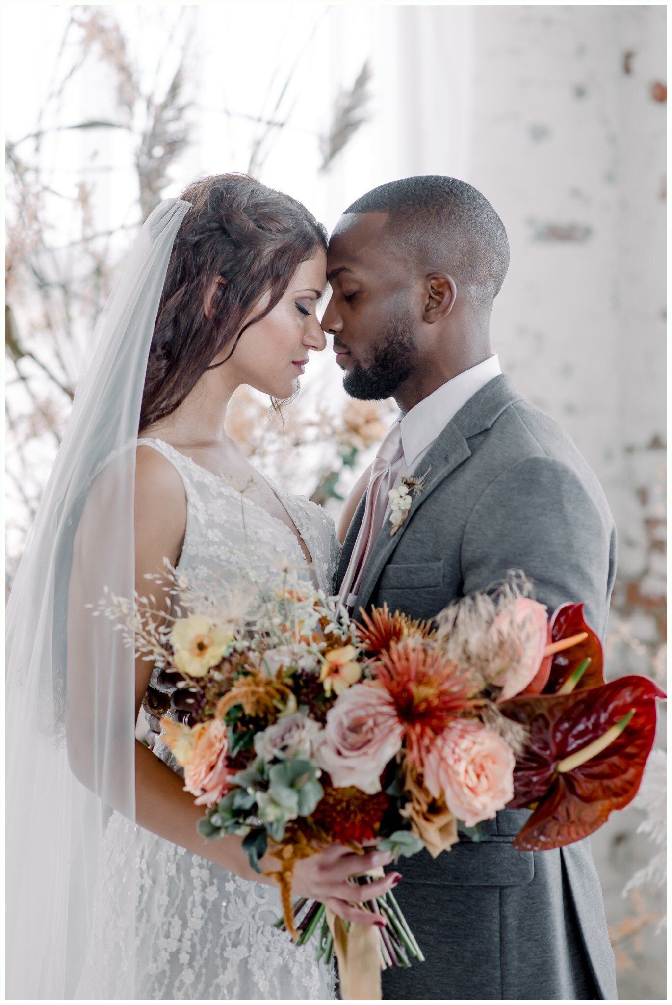 Bride and groom in formal wear, foreheads touching, holding a bouquet of pink and orange flowers