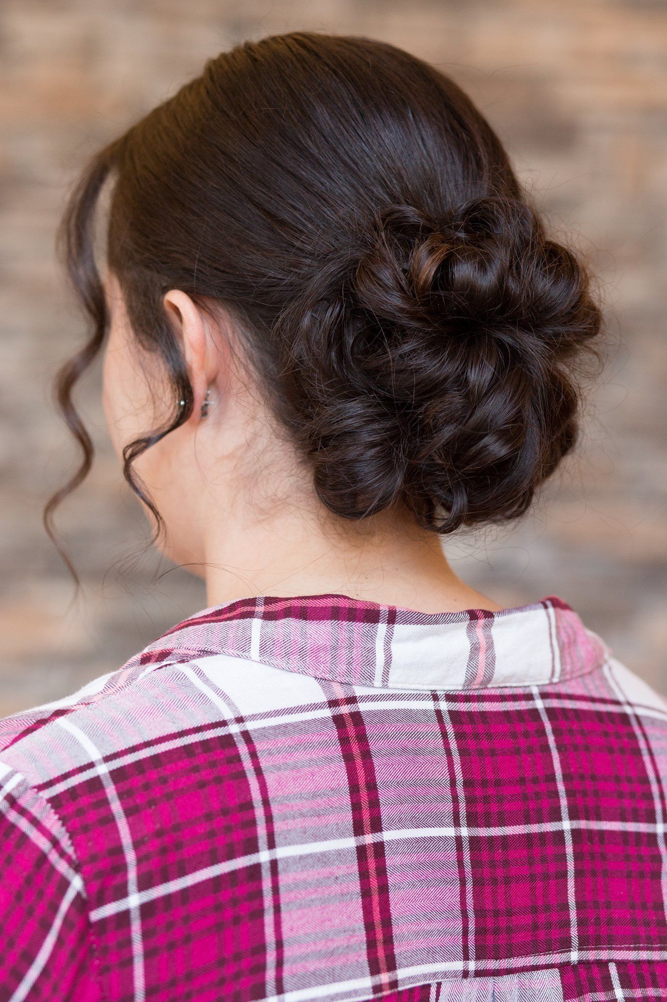 Back view of a woman with dark hair in a bun, wearing a pink plaid shirt, against a brick wall.