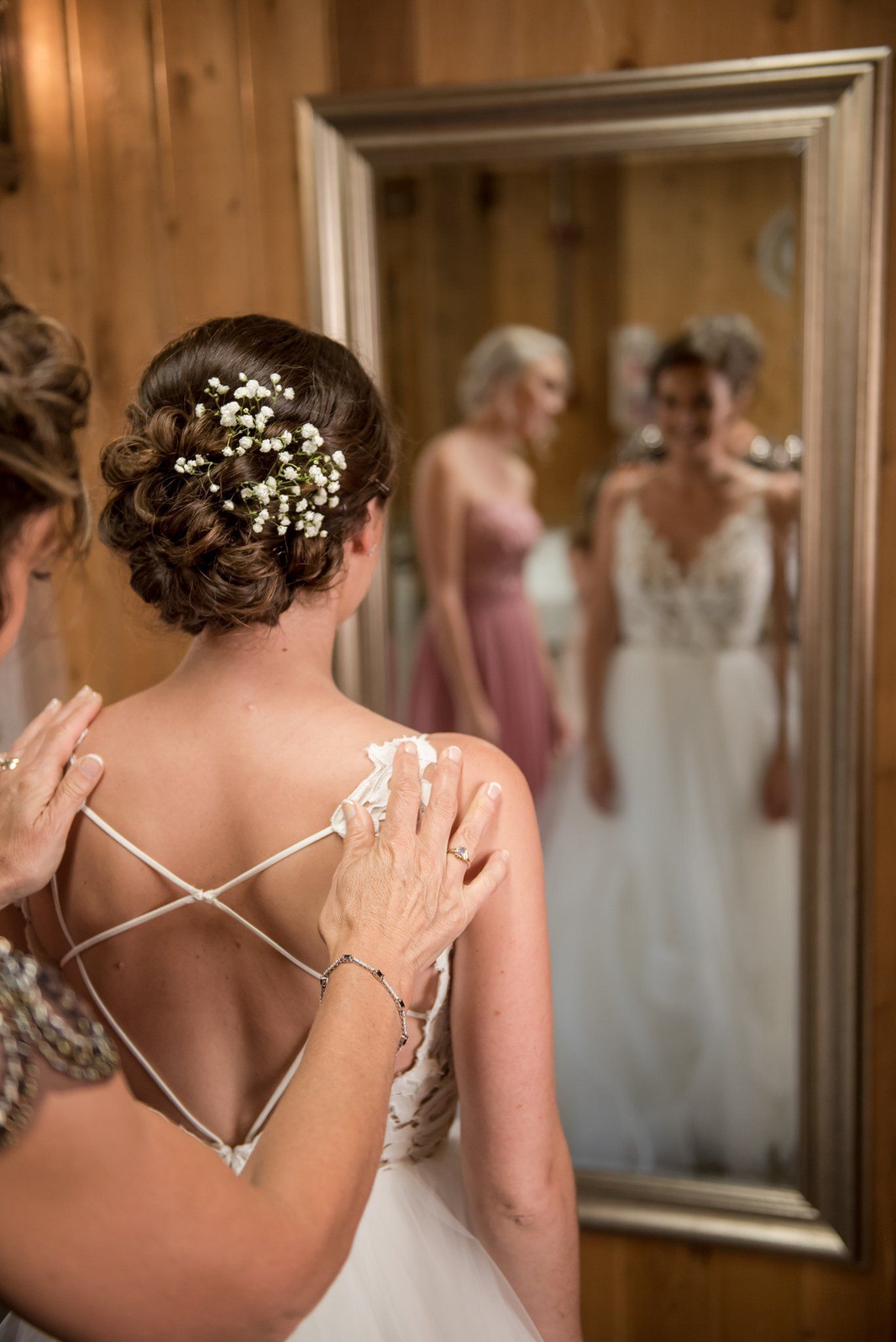 Woman in a white dress looks in a mirror while another adjusts her on her wedding day.