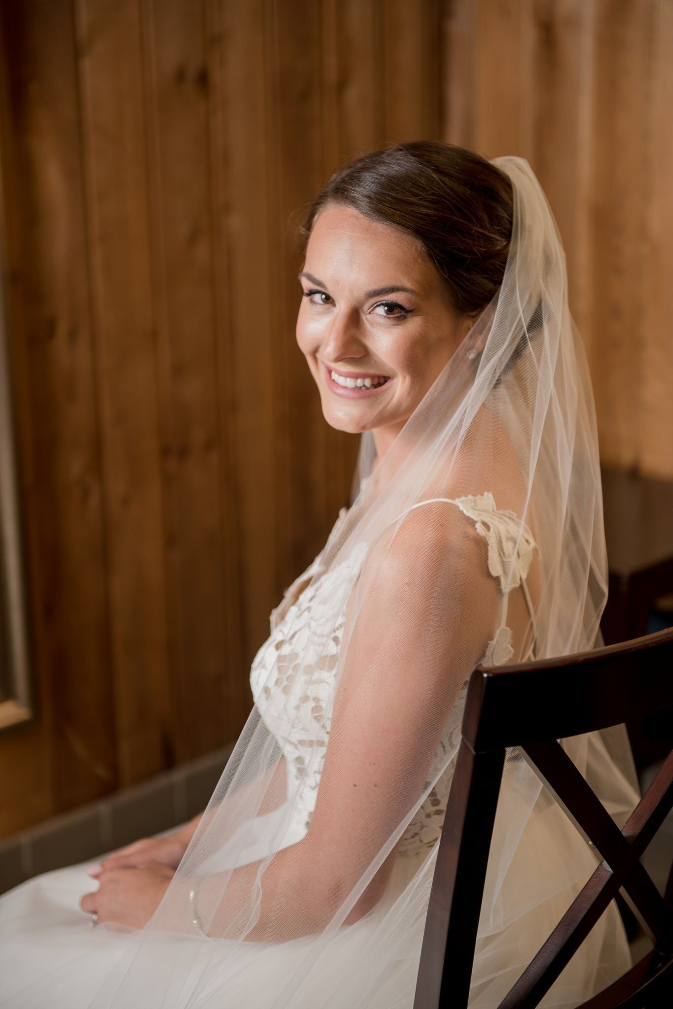 Bride in a white lace dress and veil, smiling while seated on a wooden chair indoors.