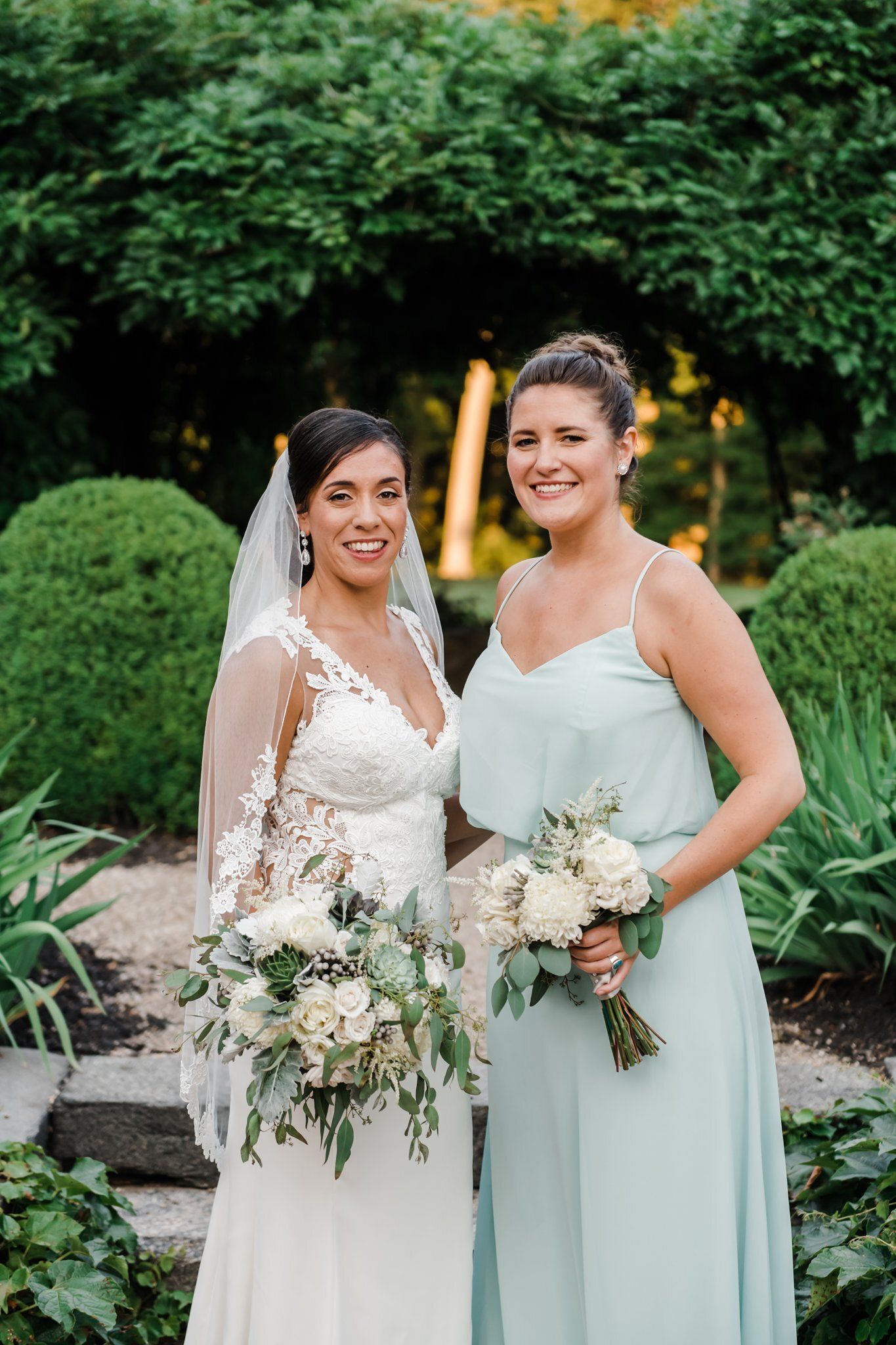 Two women in formal dresses holding bouquets, smiling in a garden setting.