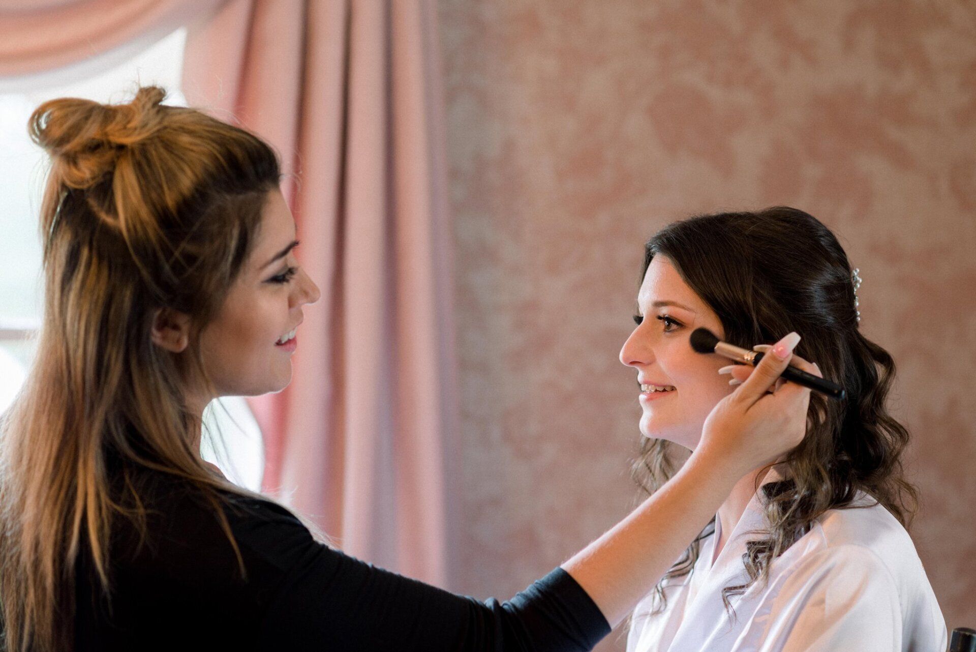 Woman applies makeup to another woman in a softly lit room.