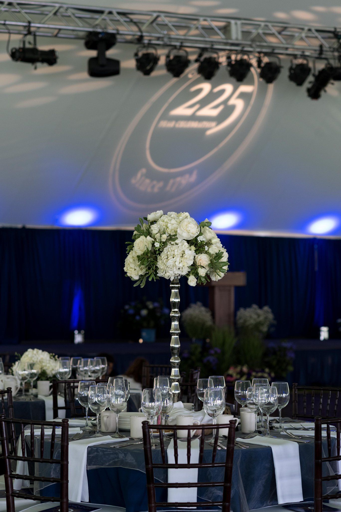 Banquet table with white floral centerpiece and silver decor beneath a stage projection of “25”