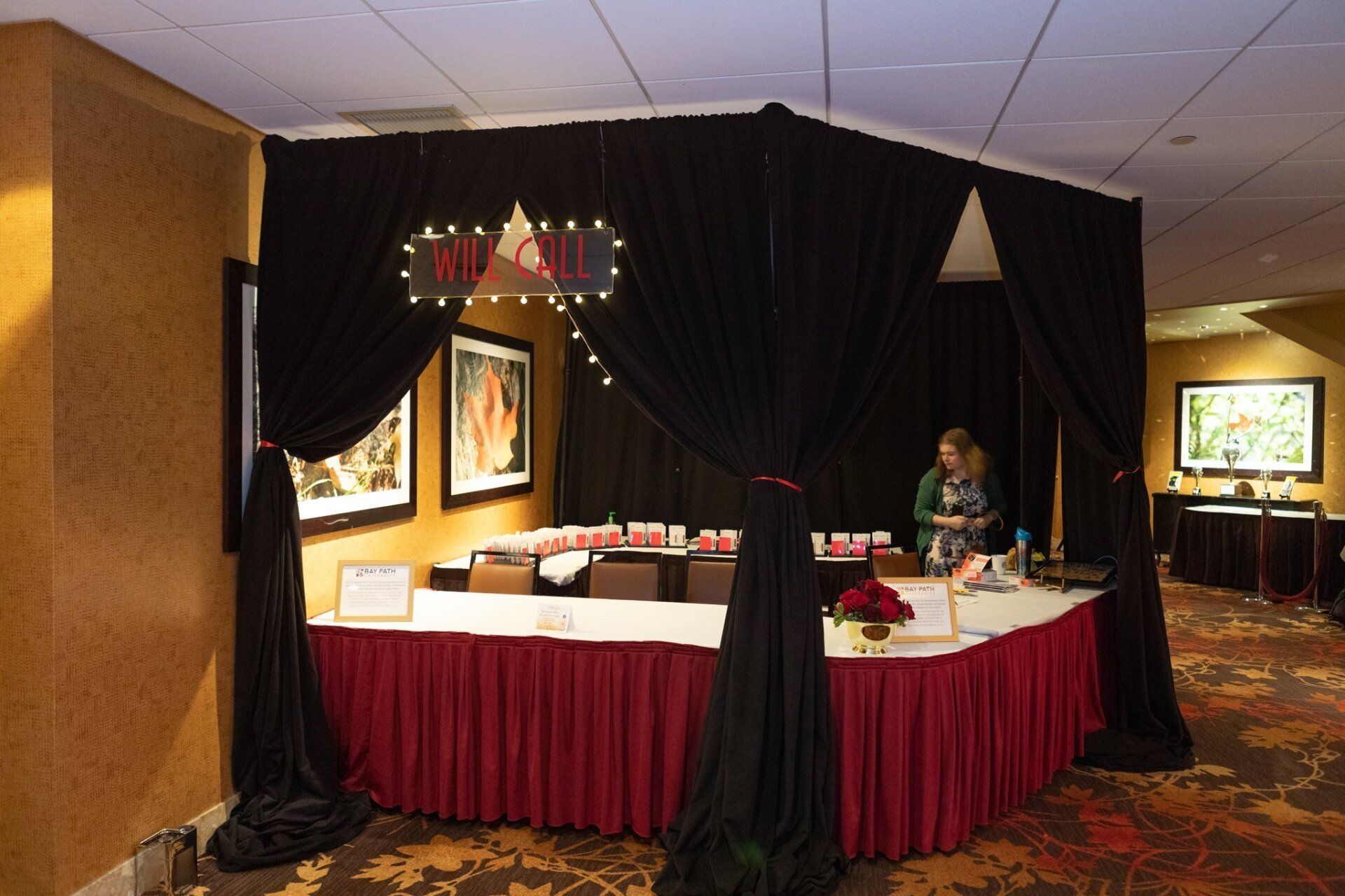 Decorated banquet booth with black drapes, red trim, and table settings at an indoor event; person stands inside.