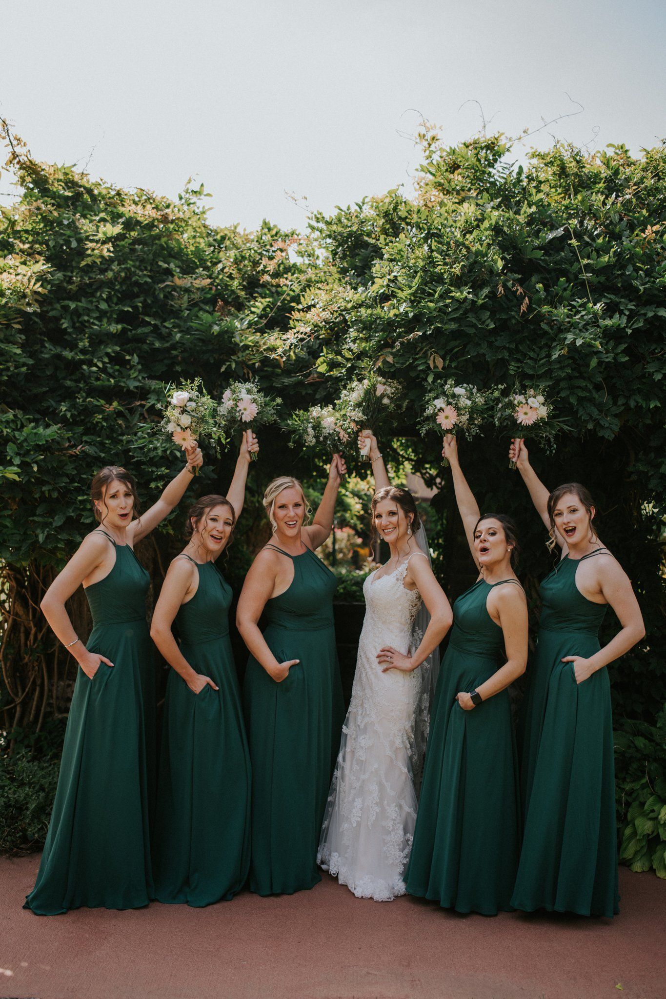 Bridal party posing outdoors, bride in white centered among bridesmaids in dark green dresses, arms raised.